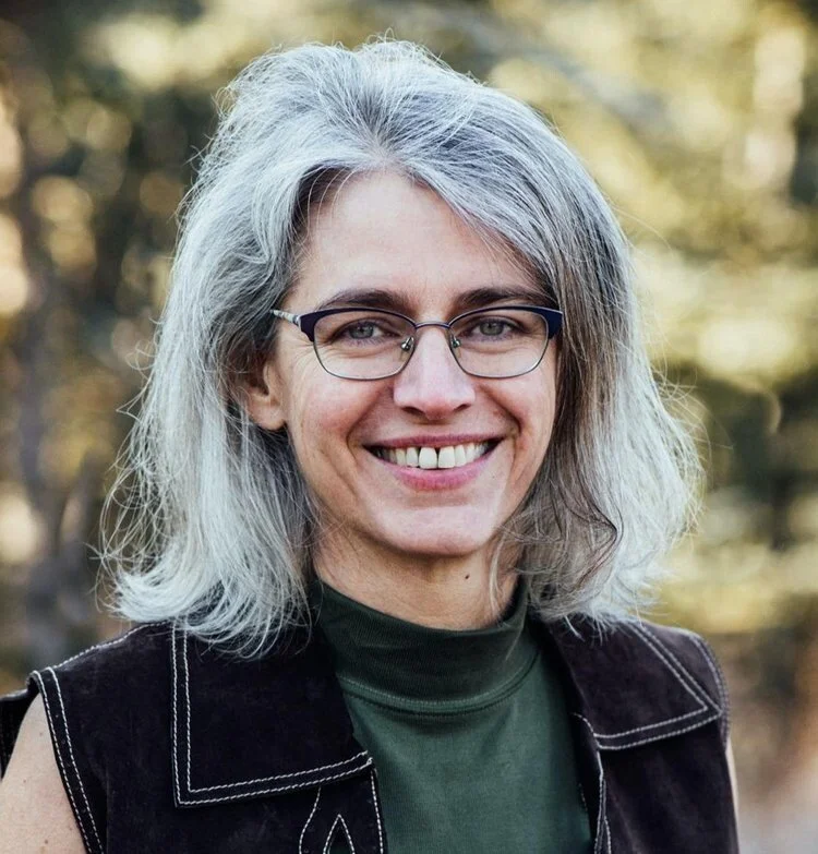 A woman with gray hair wearing glasses, smiling outdoors with blurred trees in the background.