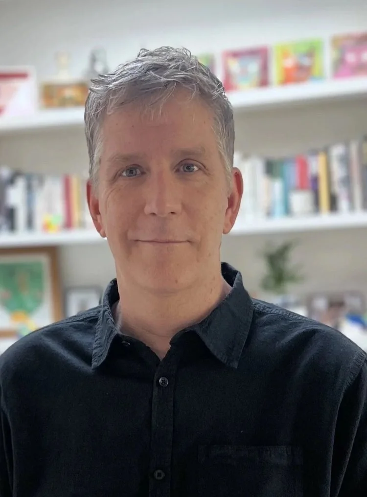 A man with short, tousled gray hair, wearing a black button-up shirt, standing in front of bookshelves filled with colorful books and decorations.