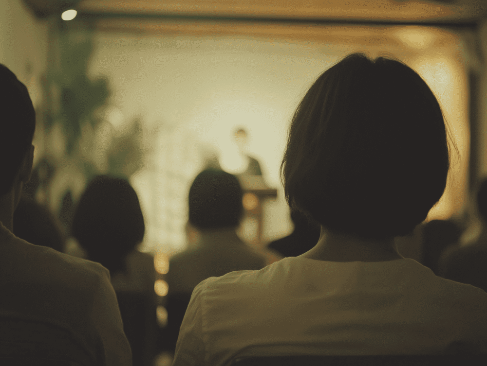 Person with short brown hair attending a presentation or lecture in a dimly lit room, with audience members visible in the background.