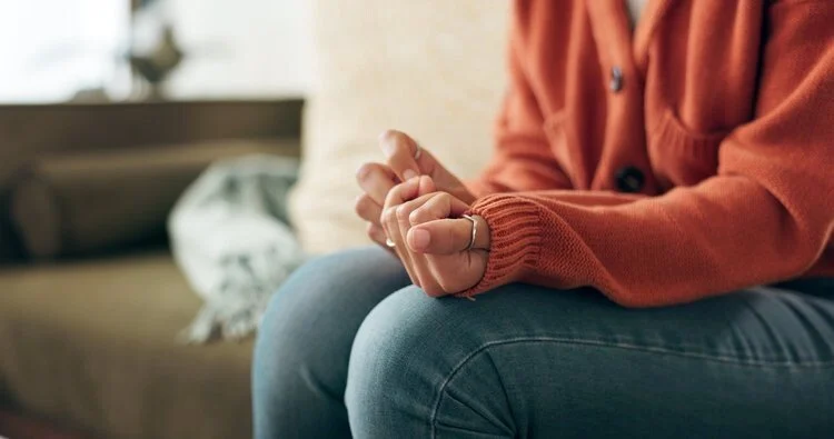 Person sitting on a couch with hands clasped, wearing an orange sweater and blue jeans.