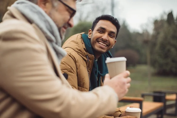 Two men sitting outdoors, smiling and holding coffee cups, with trees and benches in the background.