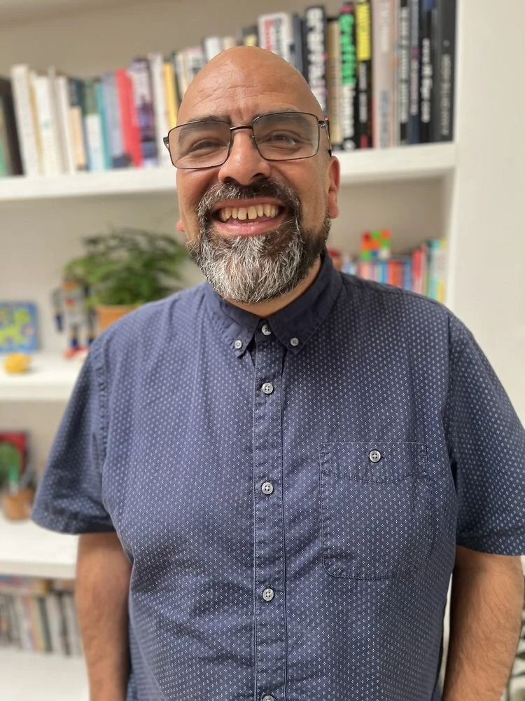 A smiling man with glasses, a beard, and a bald head, standing in front of a bookshelf filled with various books and a potted plant.
