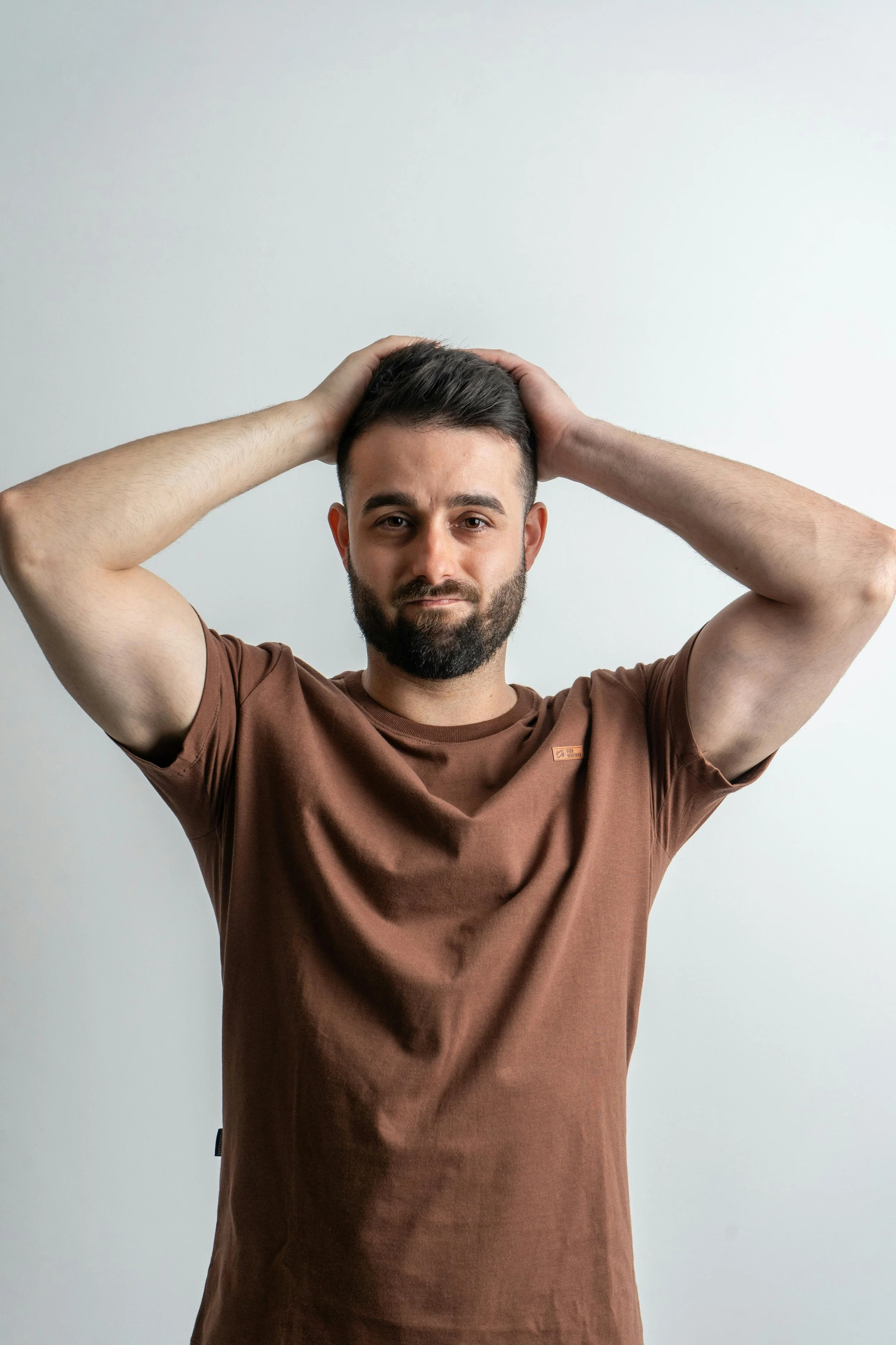 A man with a beard and short dark hair posing with his hands on his head, wearing a brown t-shirt against a plain light gray background.