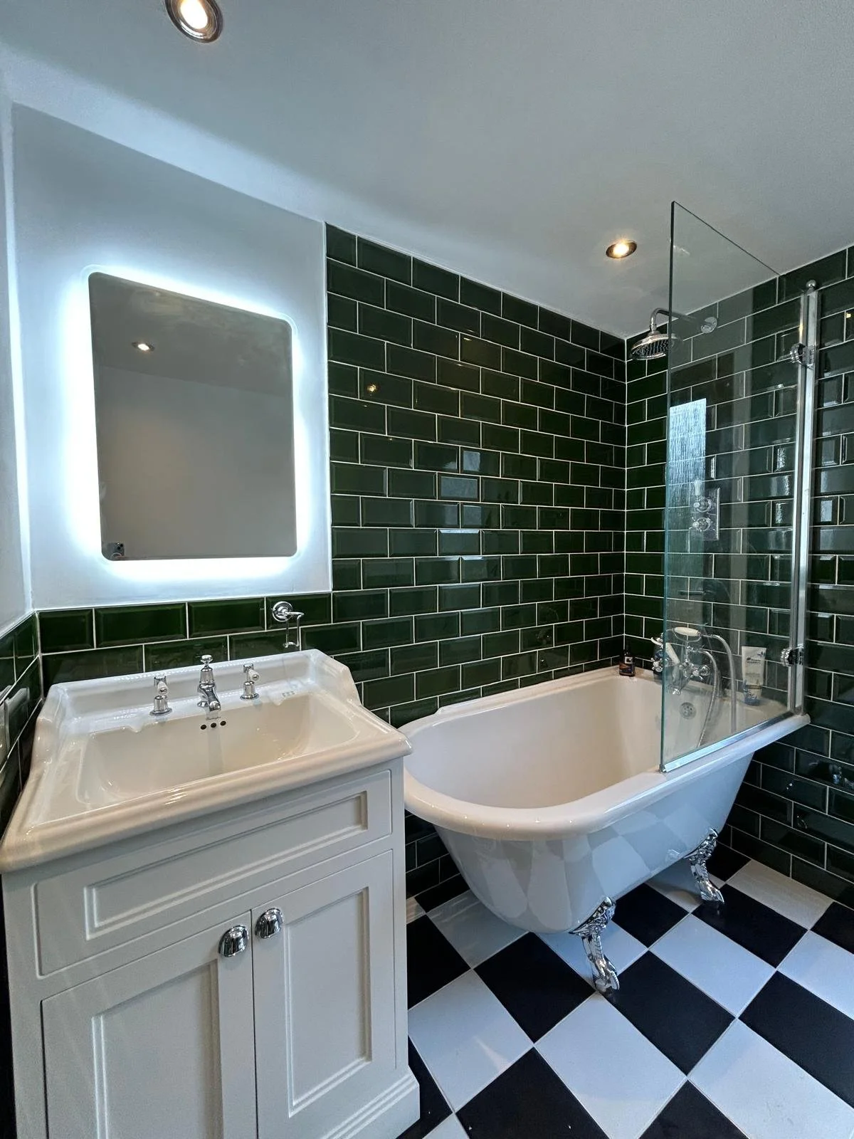 Bathroom with green subway tile walls, vintage clawfoot bathtub with decorative legs, glass shower partition, white vanity with mirror, black and white checkered floor, recessed ceiling lights in Hackney Clapton, London.