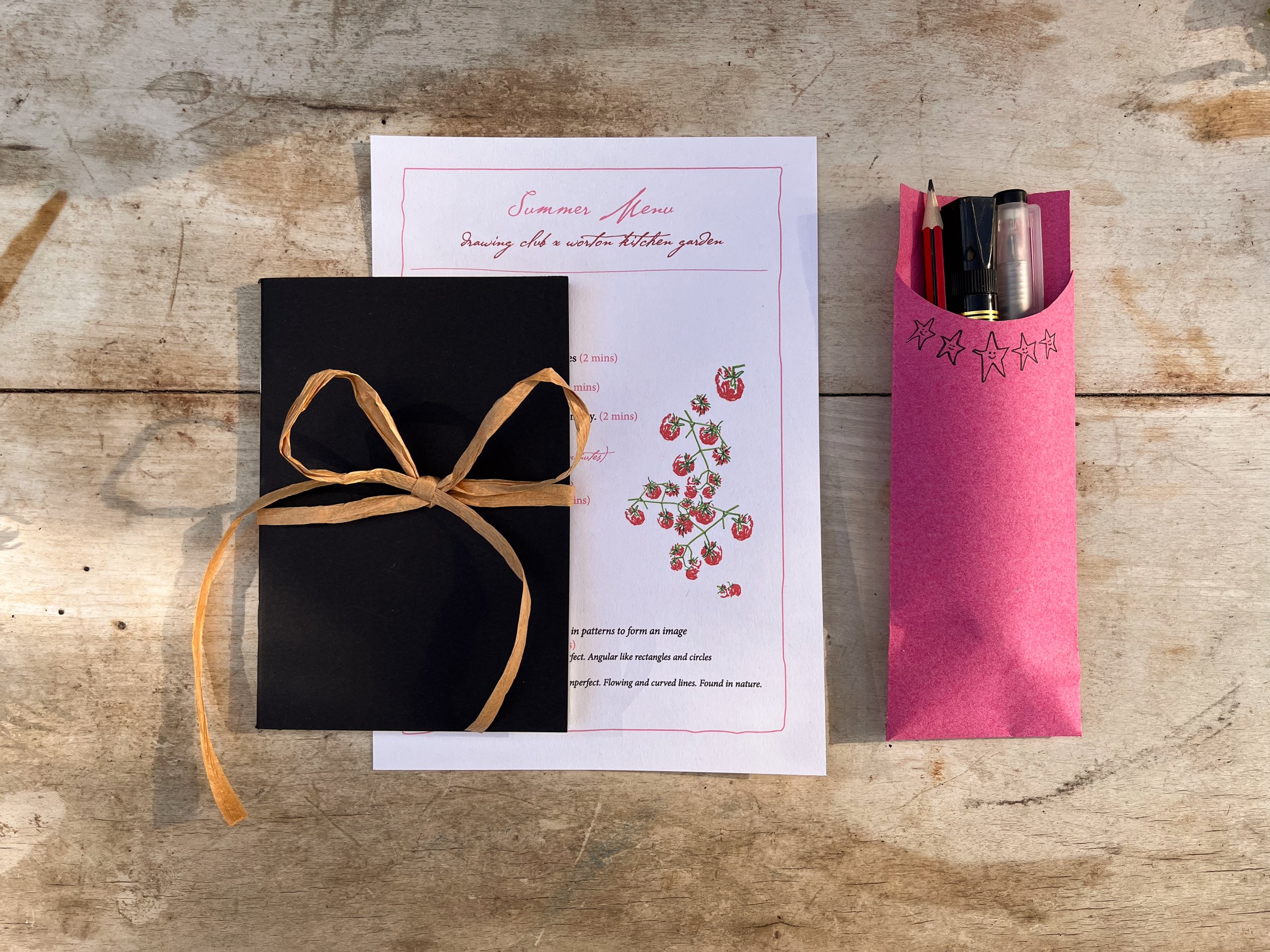 Sketchbook wrapped in a raffia bow on top of an illustrated drawing menu and pouch with pens.