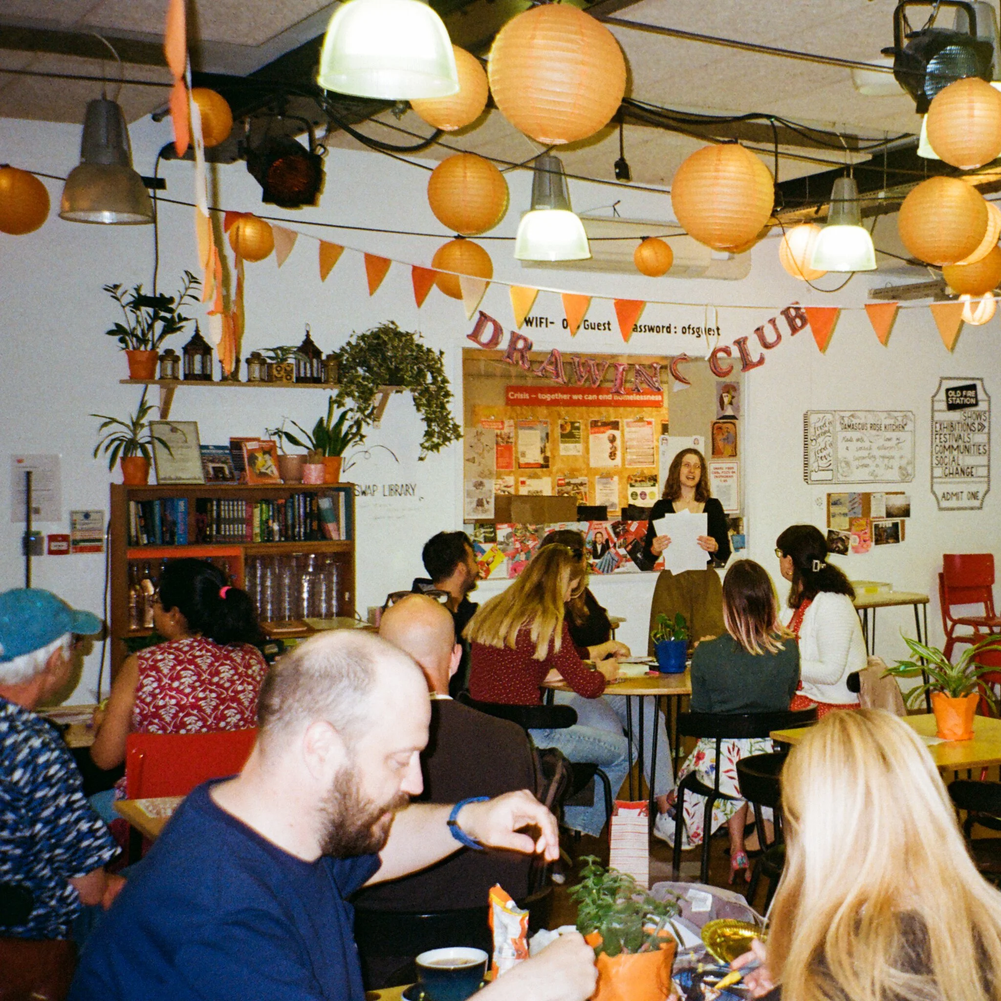 Host Bethan presenting to a full room. Orange ceiling lanterns and triangle bunting. 