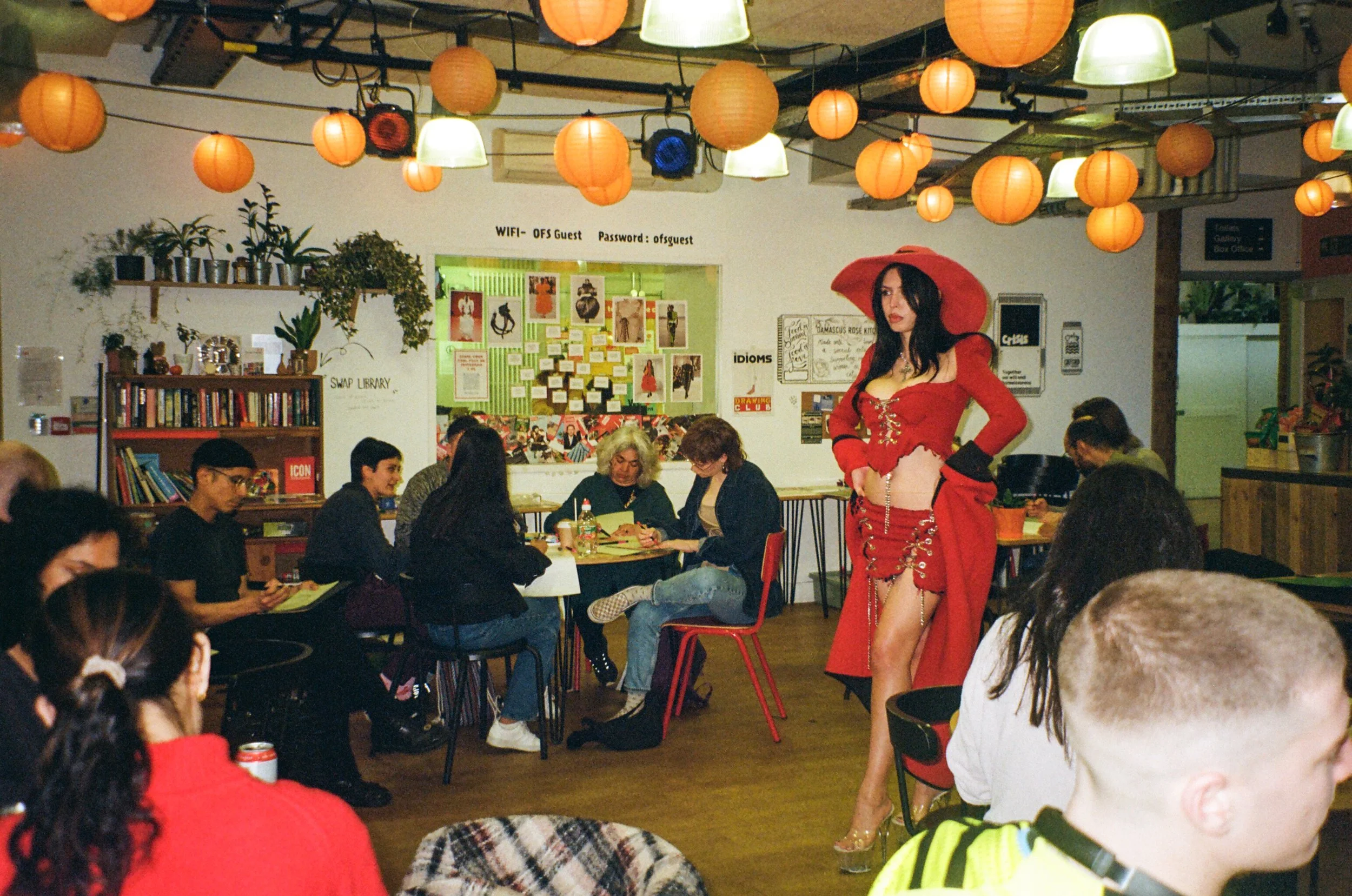 Fashion illustration model posing hands-on-hips in red hat, custom made red corset and skirt, in the middle of the room. People drawing and chatting all around her.