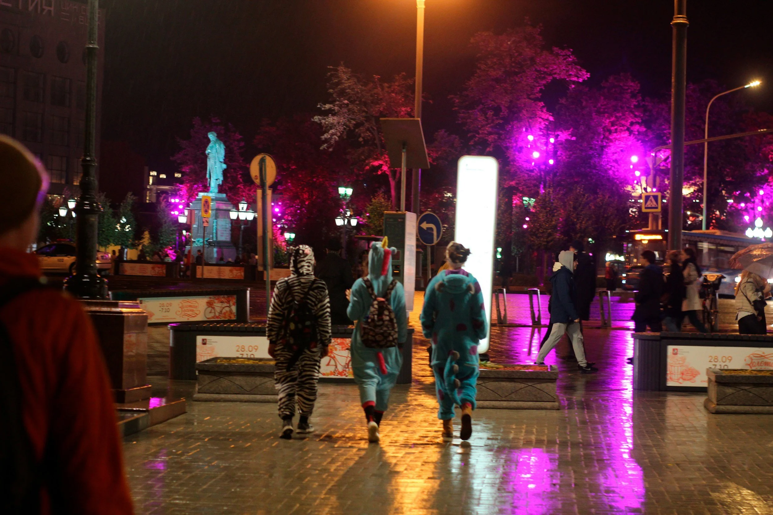 Night scene at a city street with people walking in colorful animal pajamas, illuminated trees, and streetlights reflected on the wet pavement.