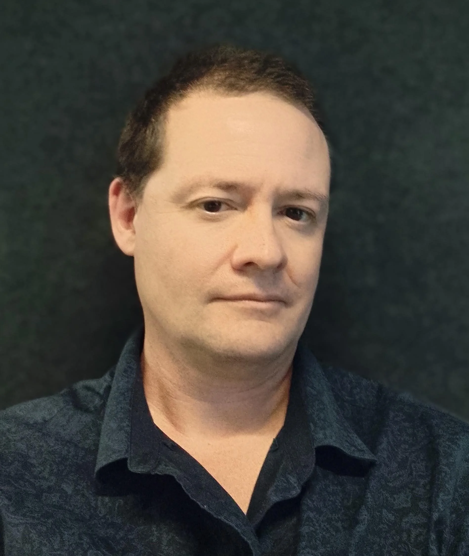 A man with short dark hair wearing a dark shirt against a dark background.