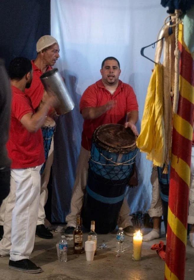 Group of men in red shirts performing a traditional musical ceremony, playing drums and a tambourine, with drinks and candles on the ground.