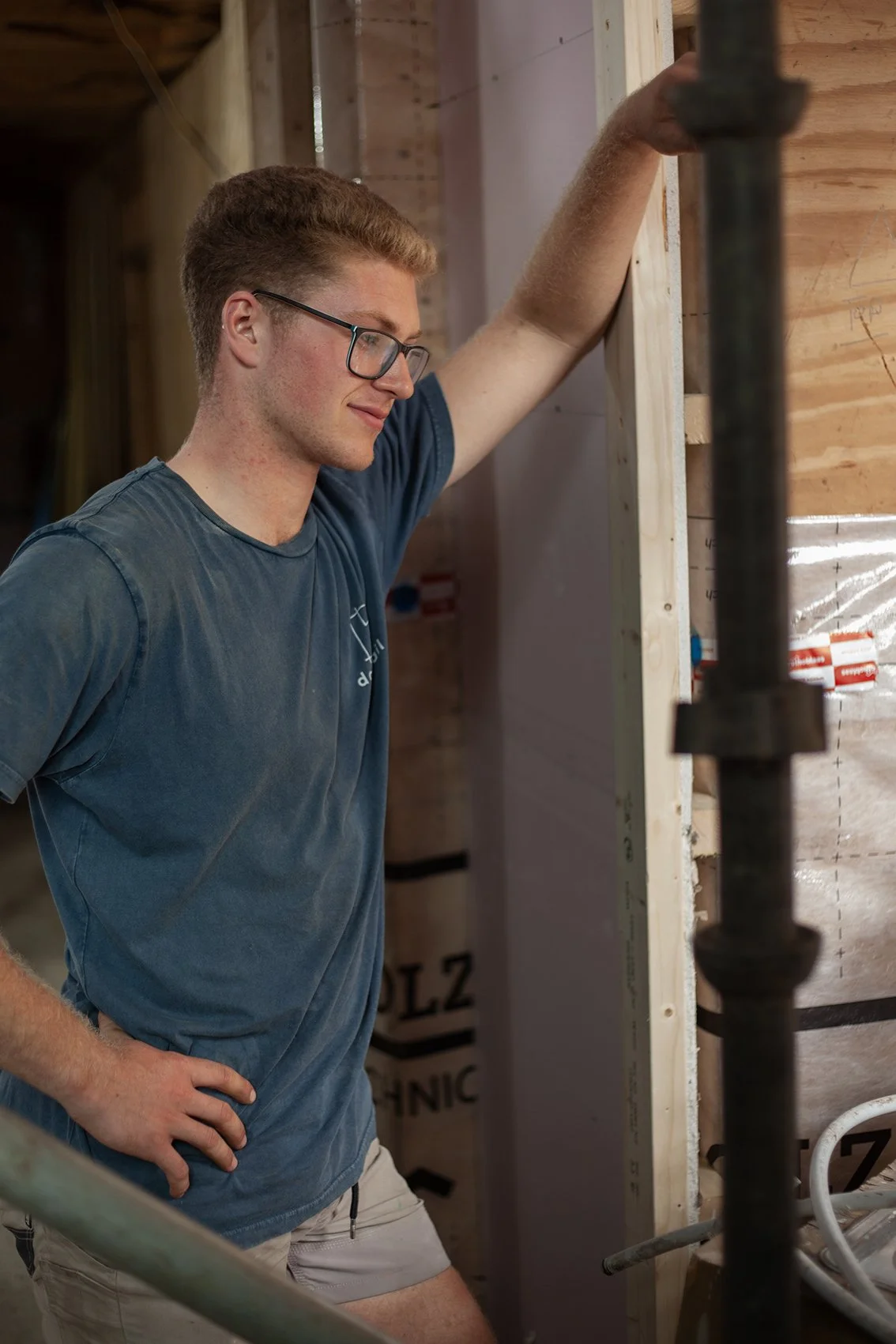 A young man with glasses stands inside a construction site, resting his arm on a wooden wall frame and looking down thoughtfully with his hand on his hip.