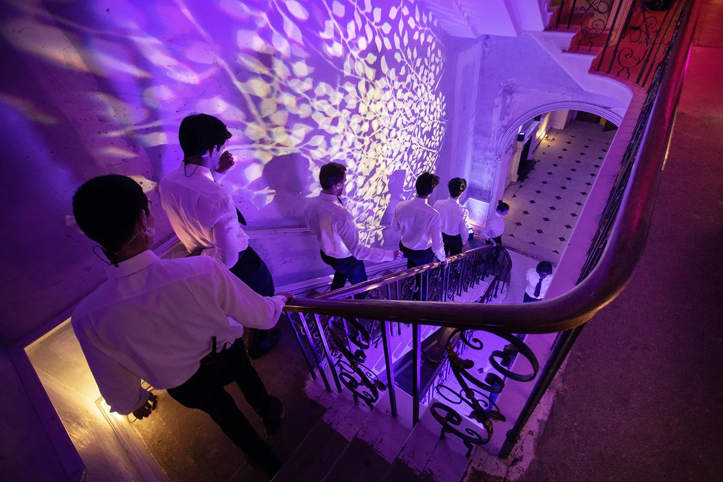A group of men dressed in white shirts and dark pants ascending a staircase in a building with ornate black railing. The wall is illuminated with purple and yellow lights creating a patterned projection. The staircase curves down to a checkered tile 