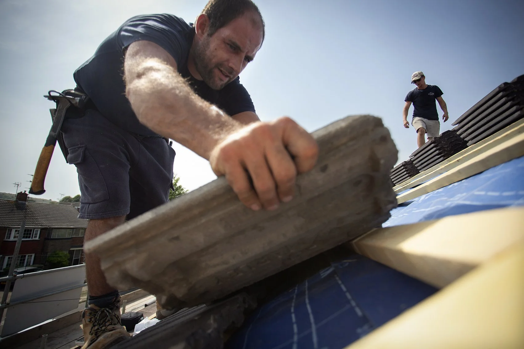 Two men working on a roof, one placing roofing tiles and the other walking on the roof. The man in the foreground is focused on the task, wearing work gear and holding a wooden board.