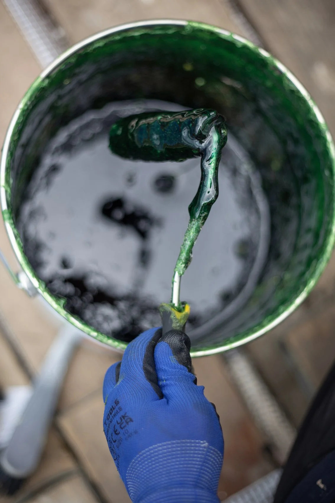 A person wearing a blue glove holding a paint roller covered in green paint over a bucket filled with green paint.
