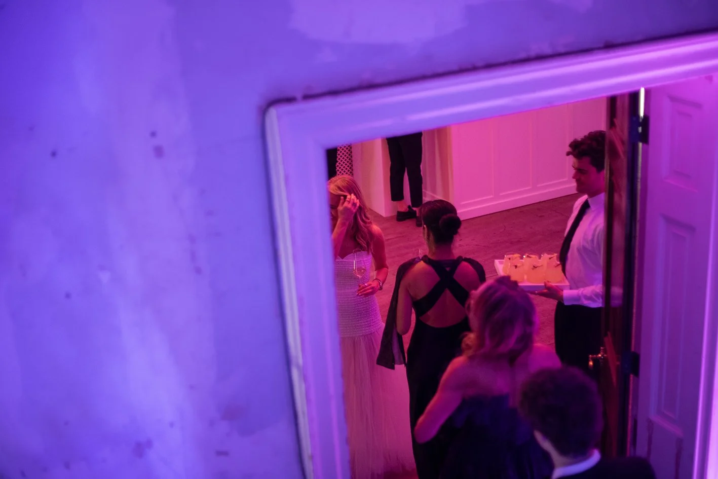 People at a party seen through a doorway, with purple and pink lighting, some holding drinks, in formal attire.