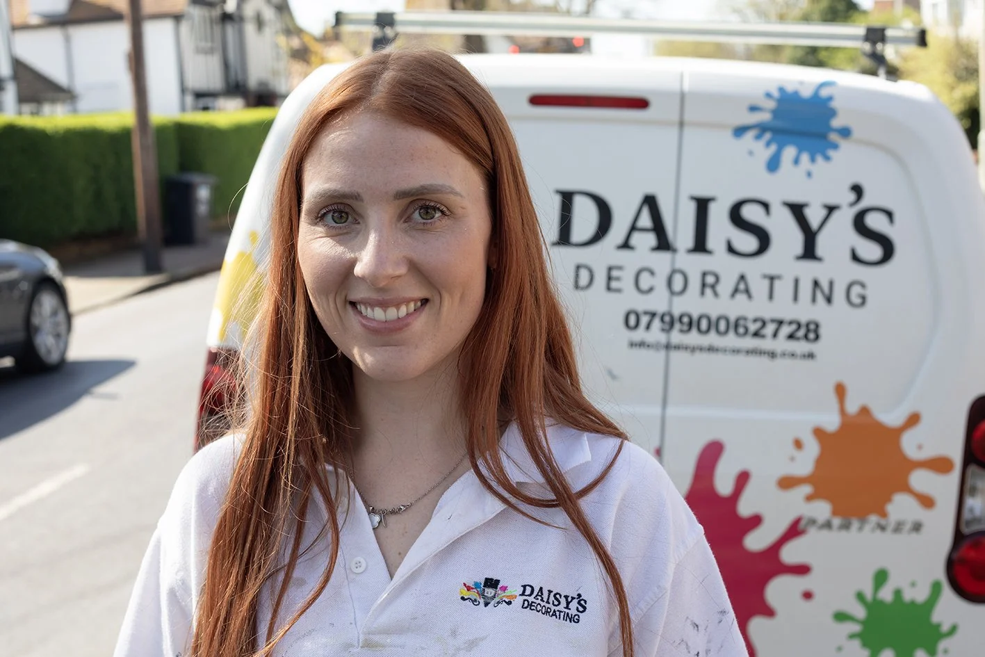 A smiling woman with long red hair wearing a white polo shirt with 'Daisy's Decorating' logo stands outdoors near a decorated van. The van has colorful paint splashes and the company's contact information.