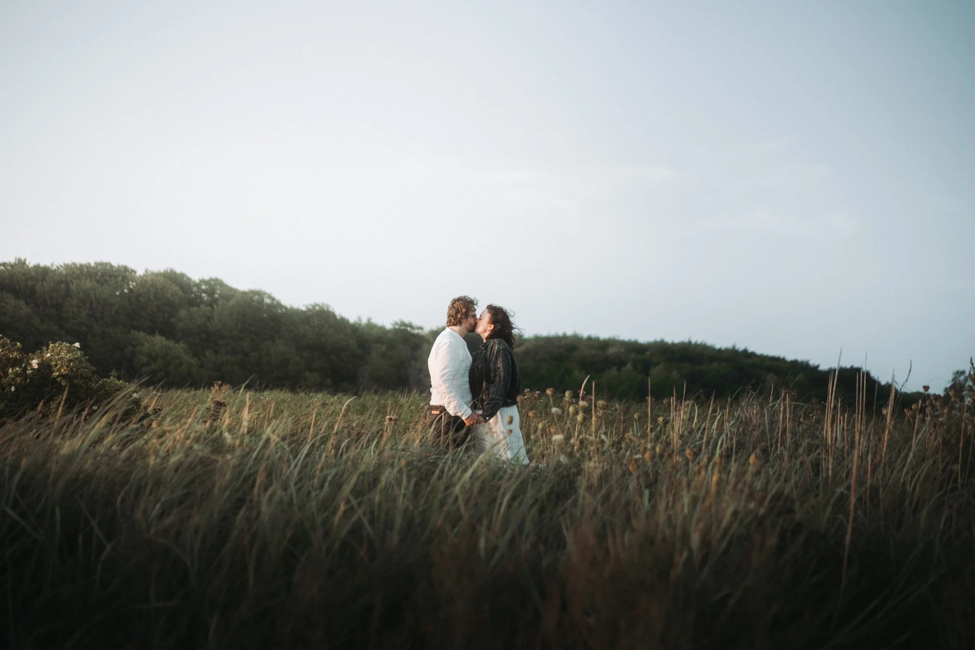 Ein Paar küsst sich im Freien, umgeben von hohen Gräsern und Bäumen im Hintergrund, bei Sonnenuntergang.