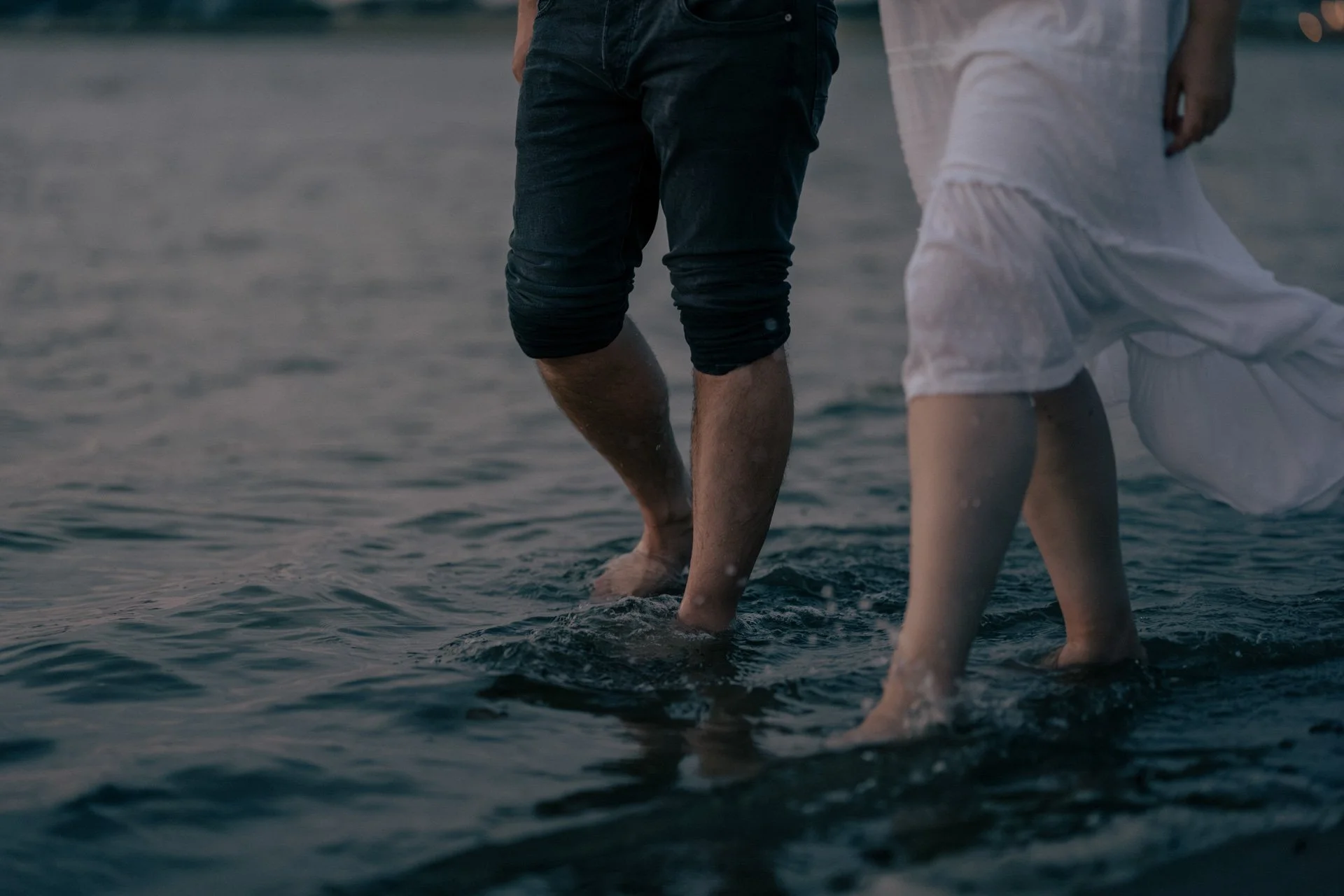 Zwei Personen, deren Füße im Wasser stehen, beim Spaziergang am Strand bei Sonnenuntergang.