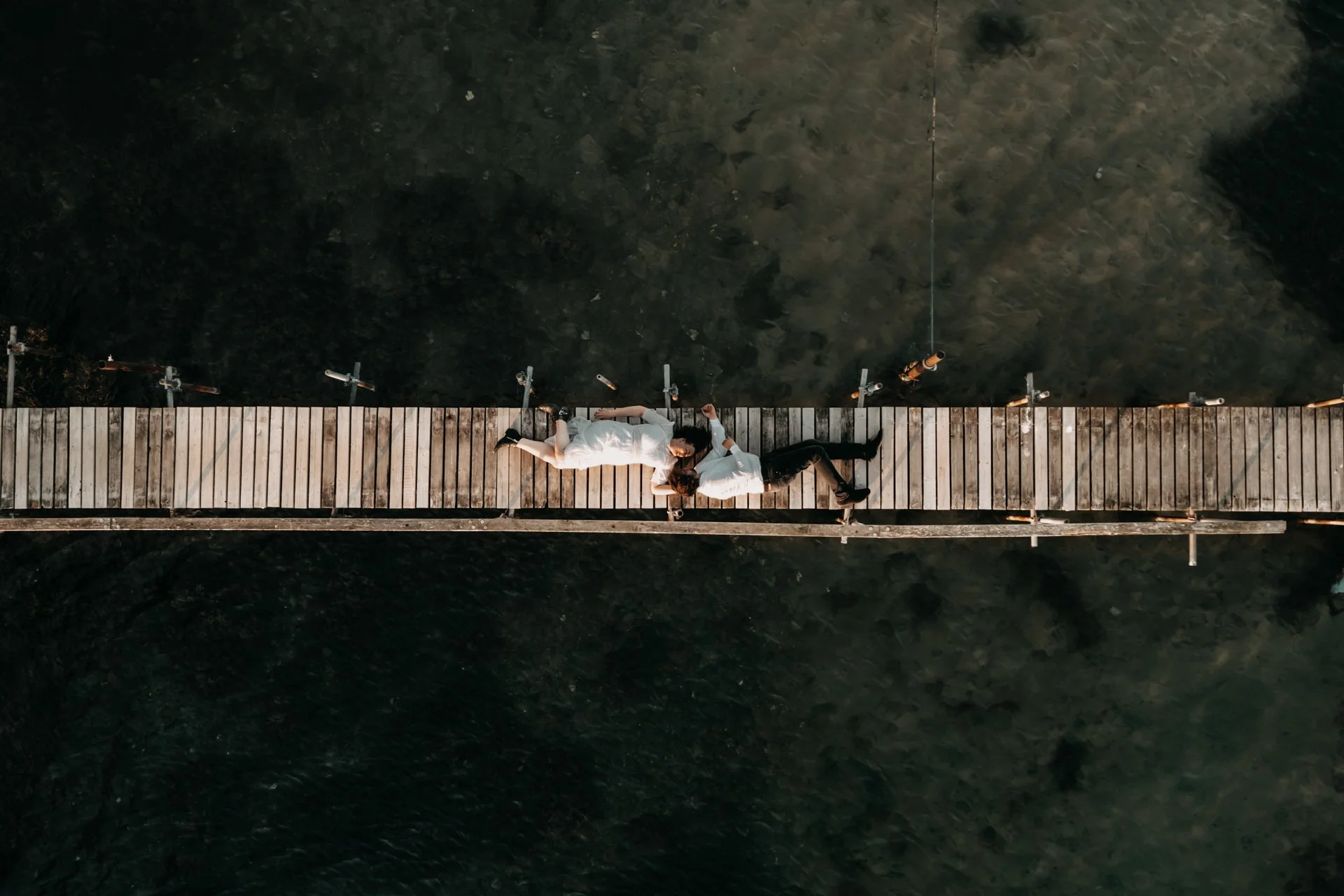 Zwei Menschen liegen auf einem kleinen Holzsteg über dunklem Wasser, die Person auf der linken trägt ein weißes Kleid, die andere schwarze Hosen, sie liegen nah beieinander und scheinen sich zu küssen.