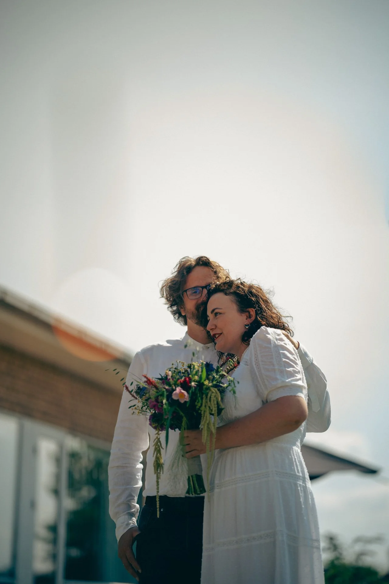 Ein Paar bei einer Hochzeit, das im Freien steht, die Frau hält einen bunten Blumenstrauß, beide lächeln und schauen sich glücklich an.