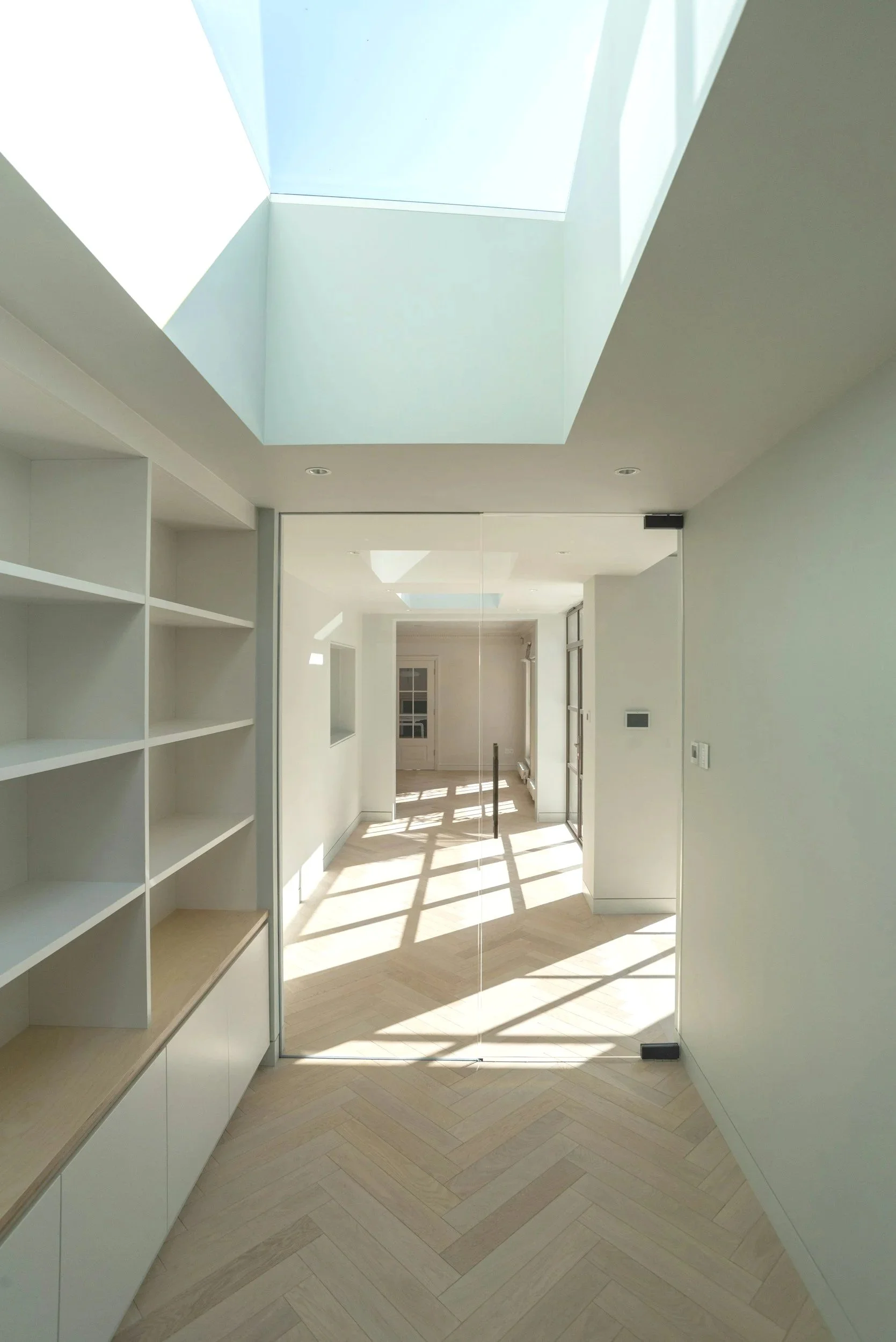 Bright modern hallway with white walls, built-in shelves on the left, a mirror sliding door, and a skylight allowing sunlight to stream in.