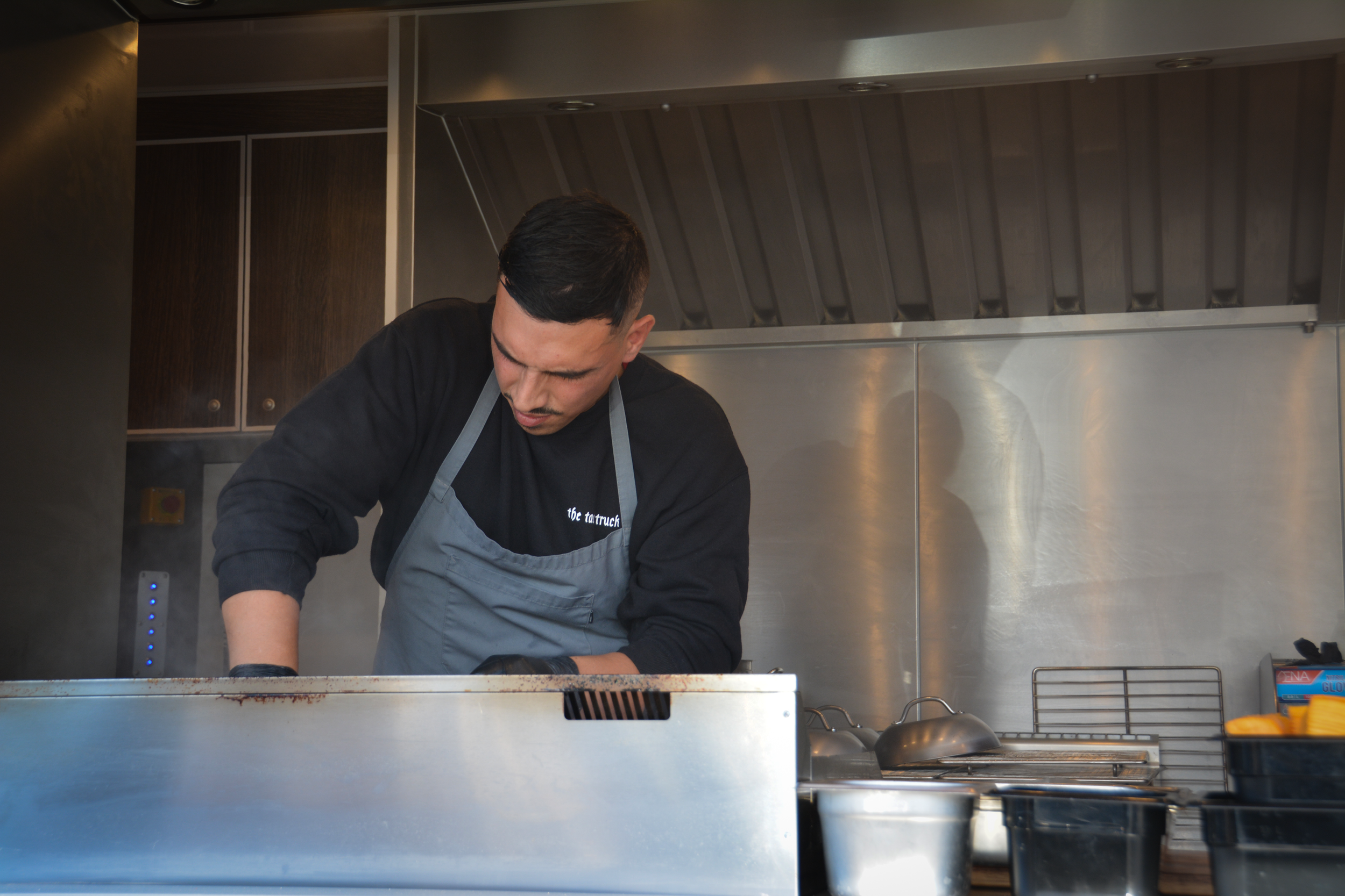 Mand, der arbejder i en professionel køkkenmiljø iført sort sweatshirt og gråt forklæde, fokuseret på madlavning │Smash Burger Truck│Copenhagen│ København