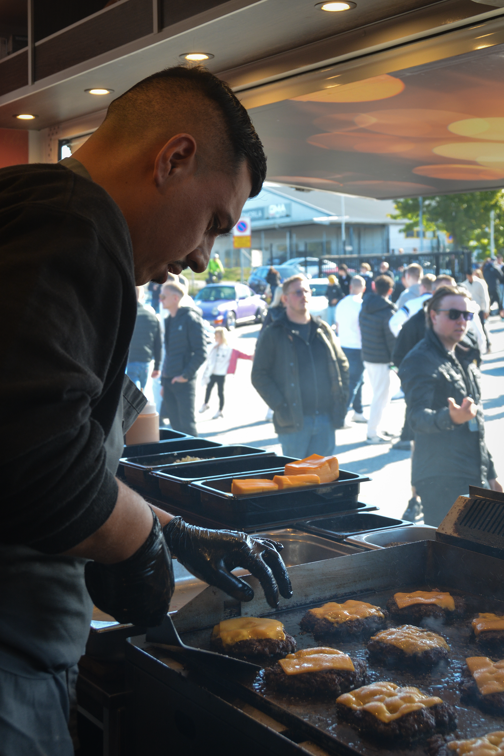 En mand tilbereder bøf med ost på en grill, mens en gruppe mennesker står udenfor i baggrunden│Smash Burger Truck│Copenhagen│ København