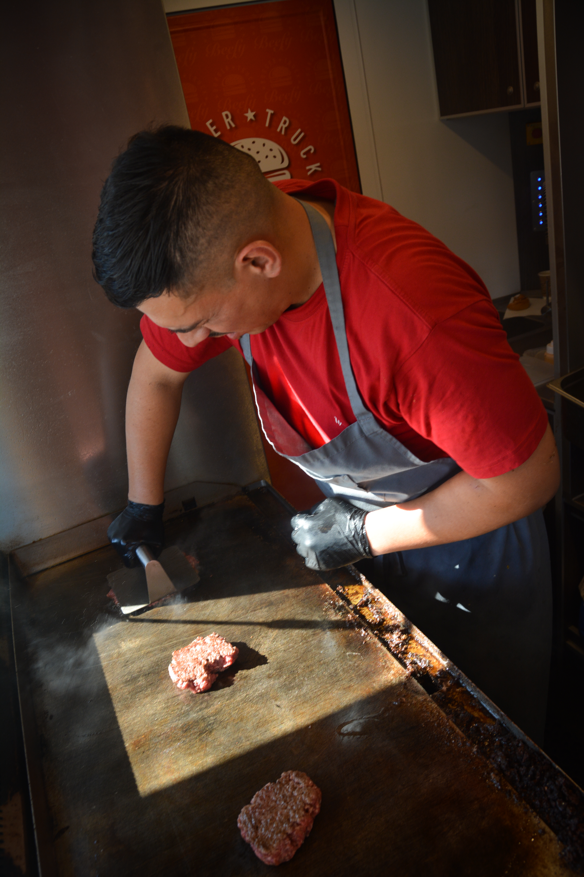 Person, der tilbereder burgerbøffer på en grillplade, iført rødt t-shirt, grå forklæde og sorte handsker │Smash Burger Truck│Copenhagen│ København