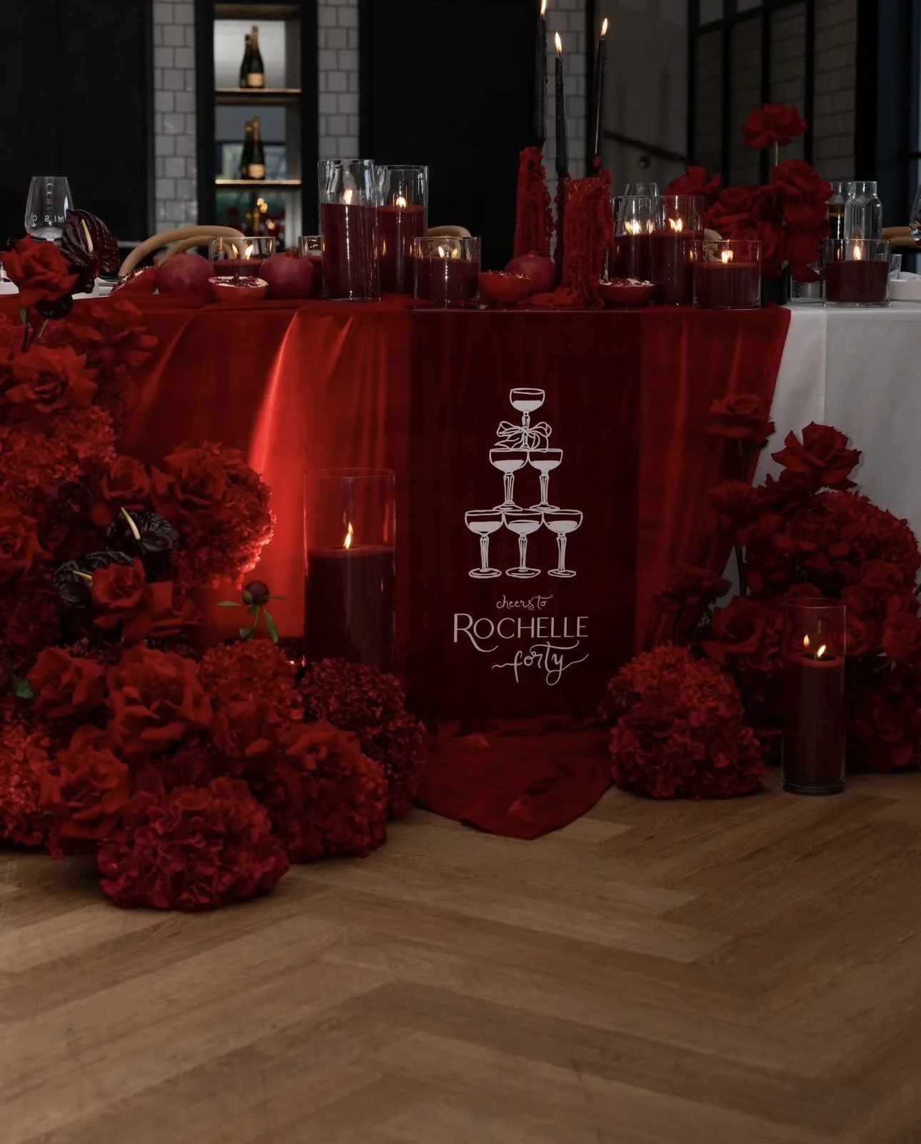 A decorated table with red flowers, candles, and a red banner with white text that reads "Cheers to Rochelle Forty."