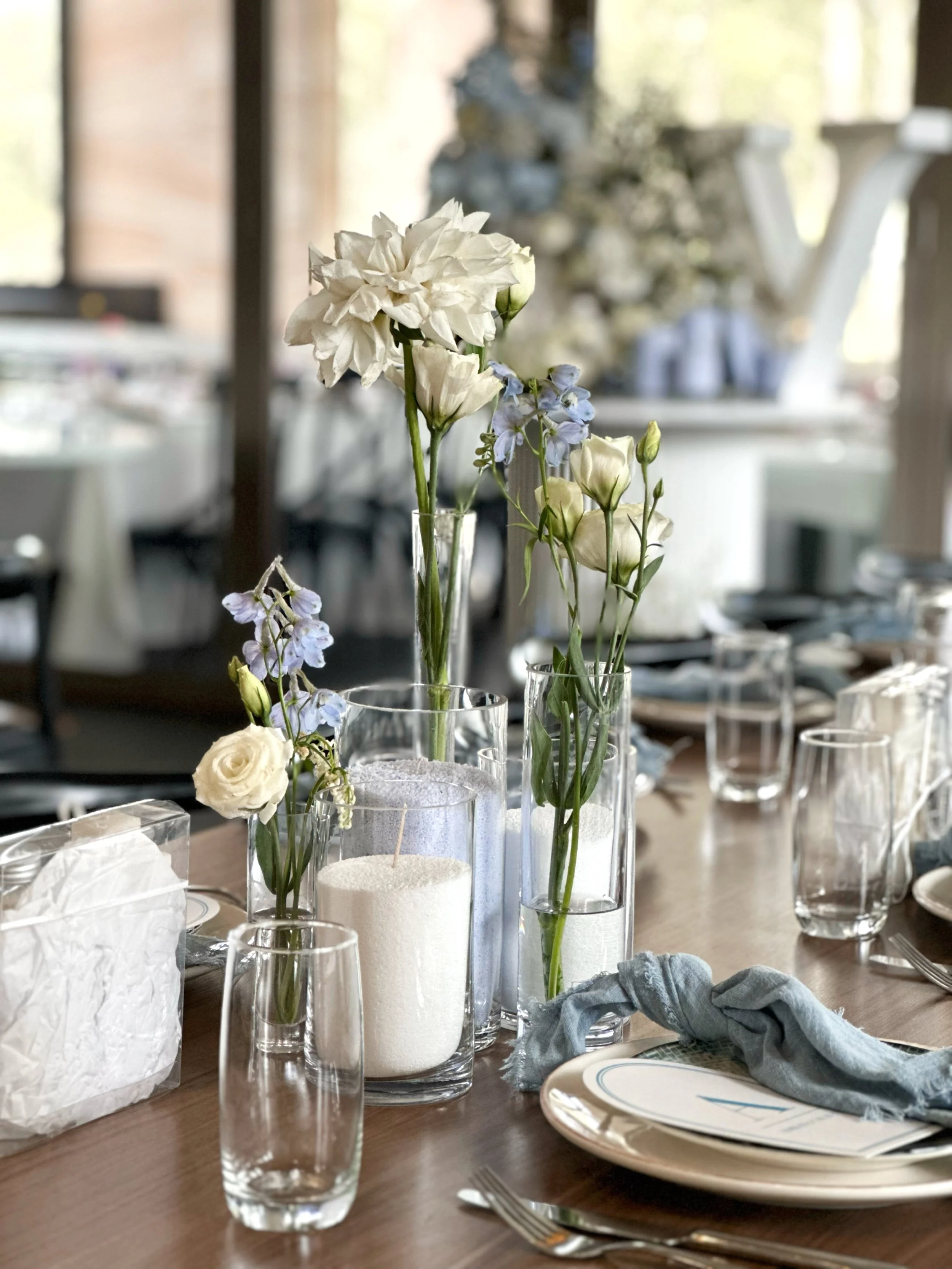 Table decorated with white flowers in glass vases, surrounded by glassware, plates, silverware, and napkins, set for a meal in a well-lit room.