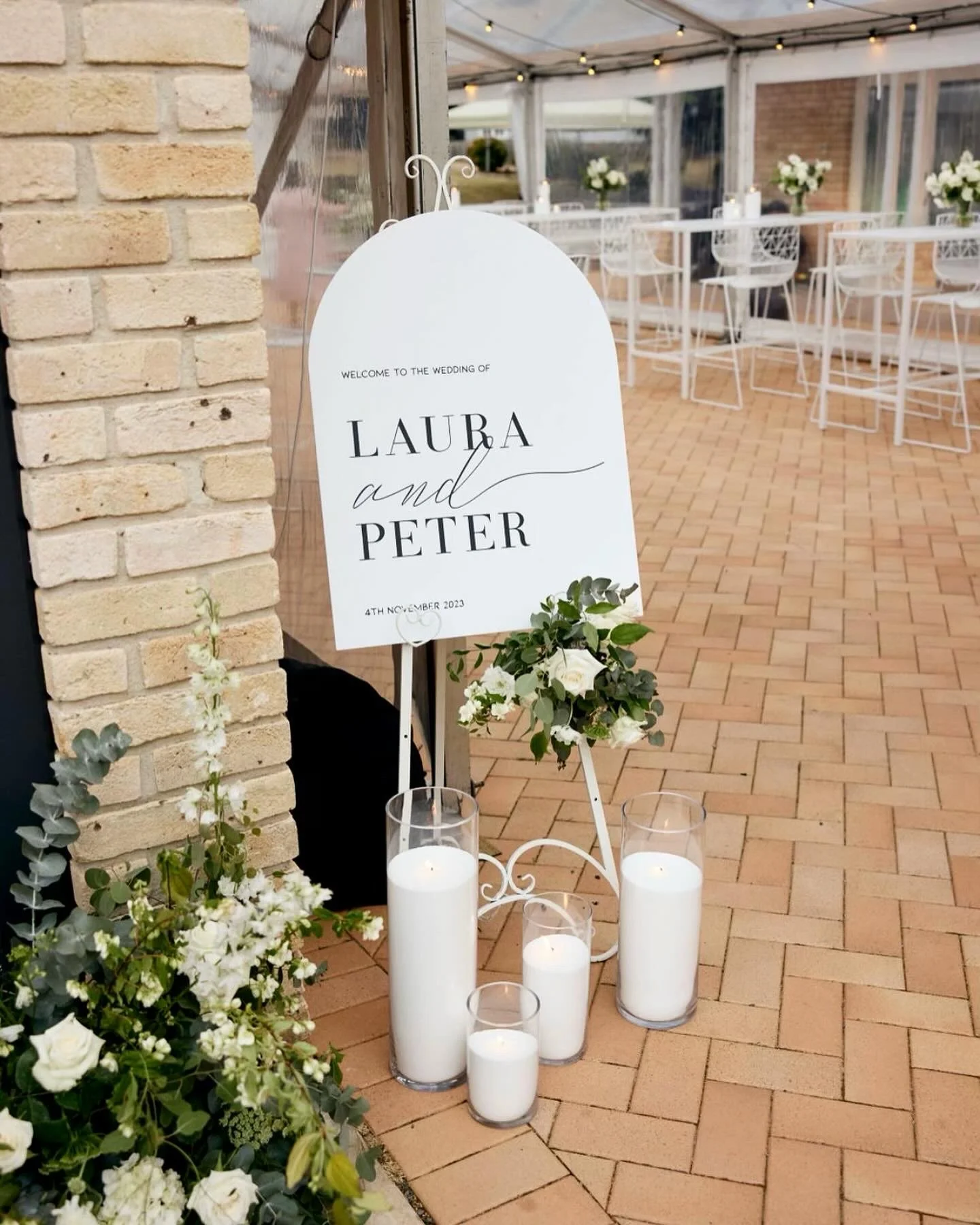 Wedding reception area with white chairs, flowers, and candles, and a sign welcoming guests to the wedding of Laura and Peter on November 4, 2023.