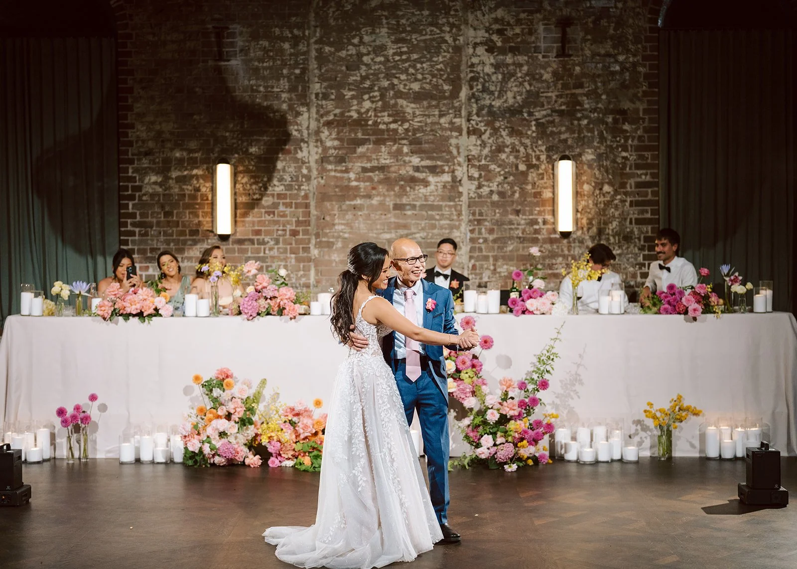 A bride and an older man, possibly her father, dance together at a wedding reception. The background shows a long table with flower arrangements, candles, and seven seated guests against a rustic brick wall.