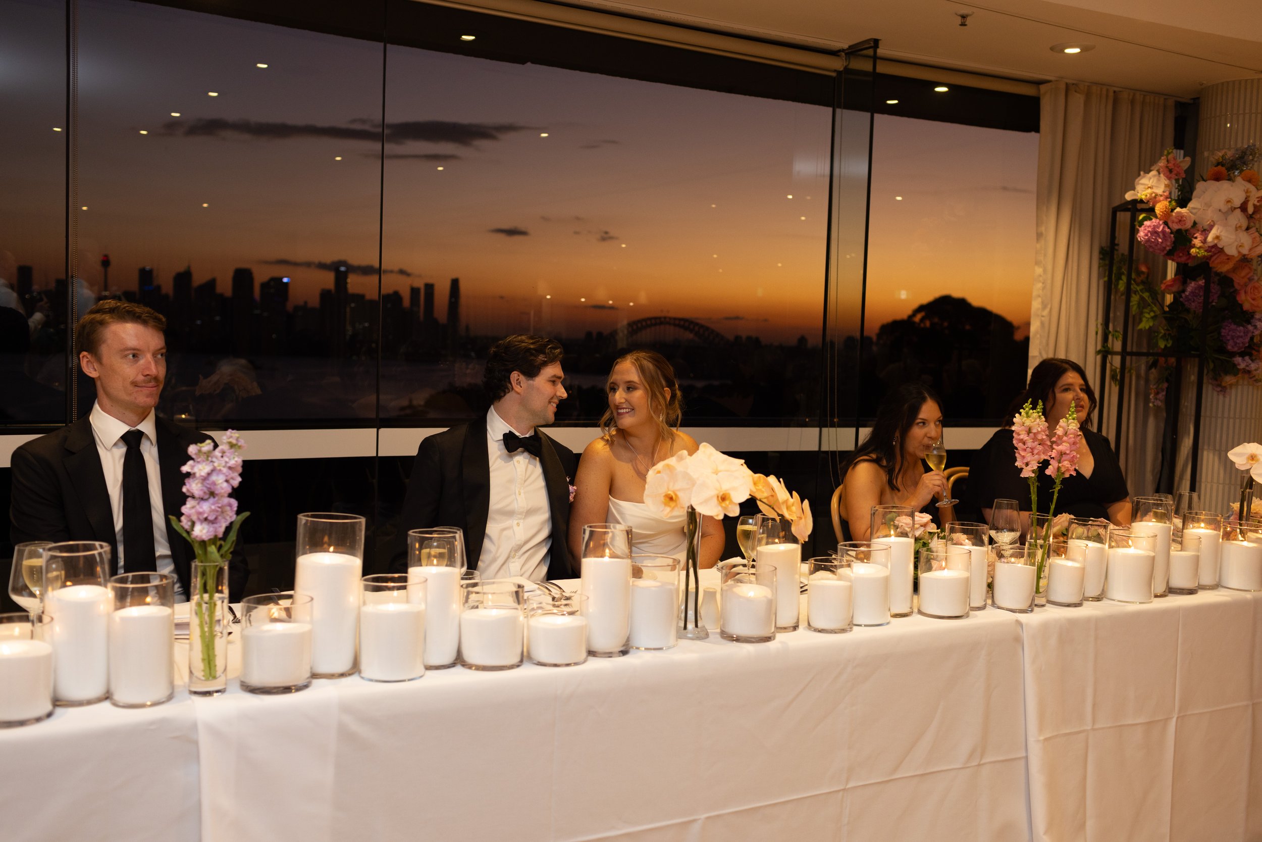 A group of wedding guests sitting at a long reception table decorated with white candles and floral arrangements, enjoying a sunset view through large windows.