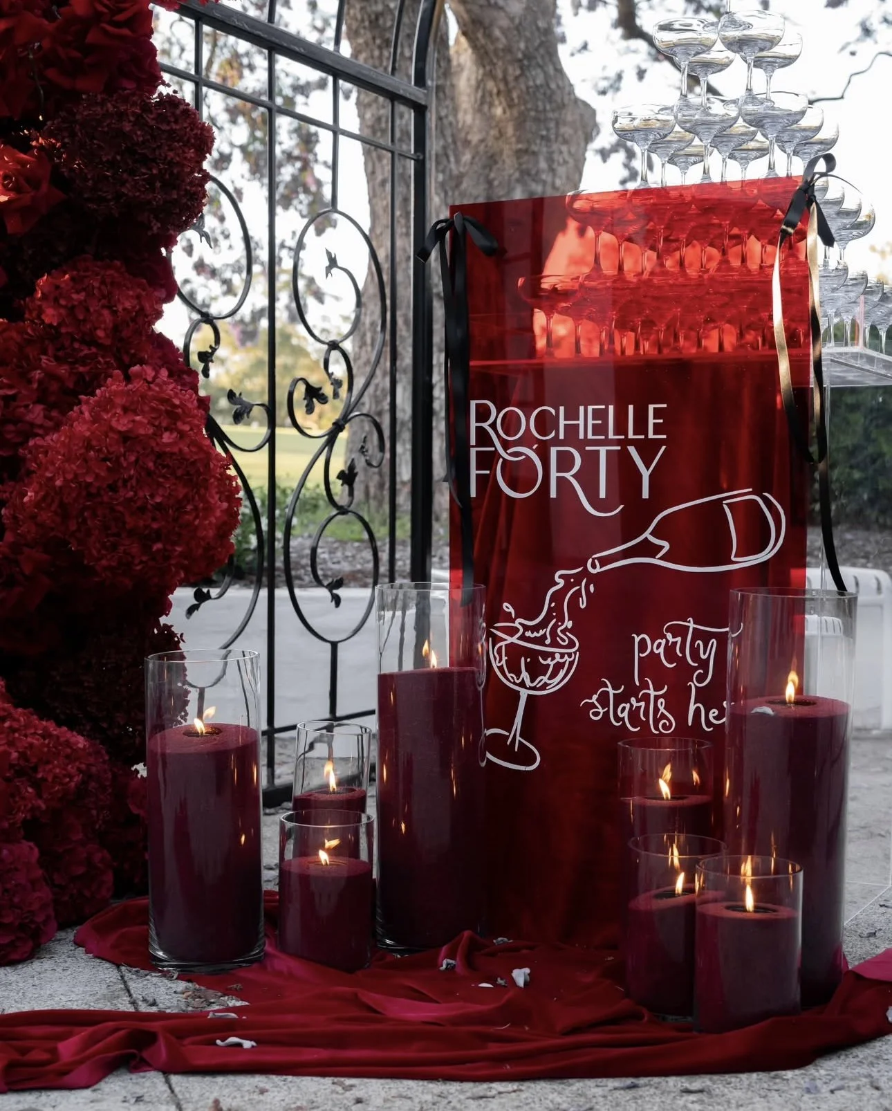 Display of a celebratory arrangement with stacked glasses, red candles, and red flowers. A red sign with the text 'Rochelle Forty' and an illustration of a champagne bottle pouring into a glass, with 'party starts here' written below.
