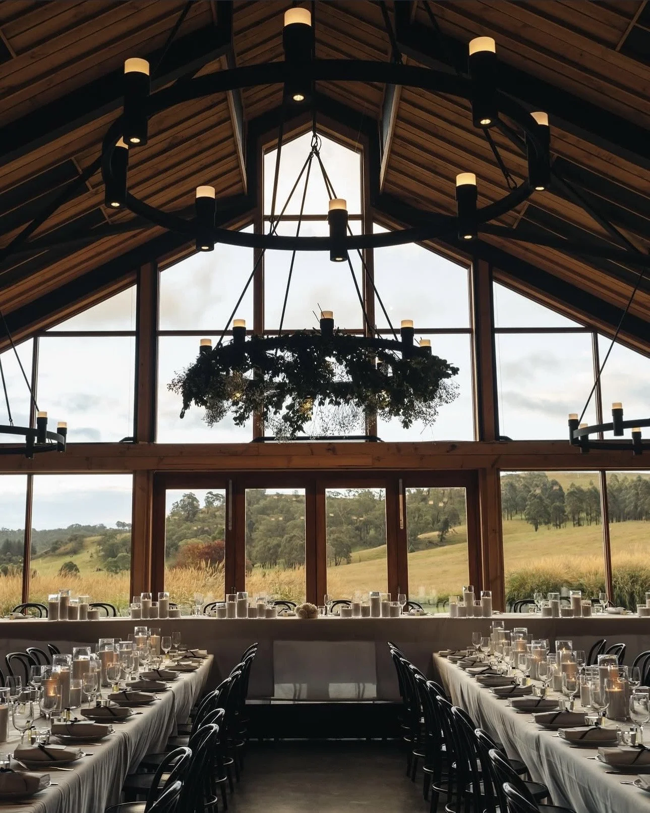 An interior view of a rustic wedding reception or banquet hall with a high wooden ceiling, large windows showing a scenic landscape, and elegant chandeliers decorated with greenery.