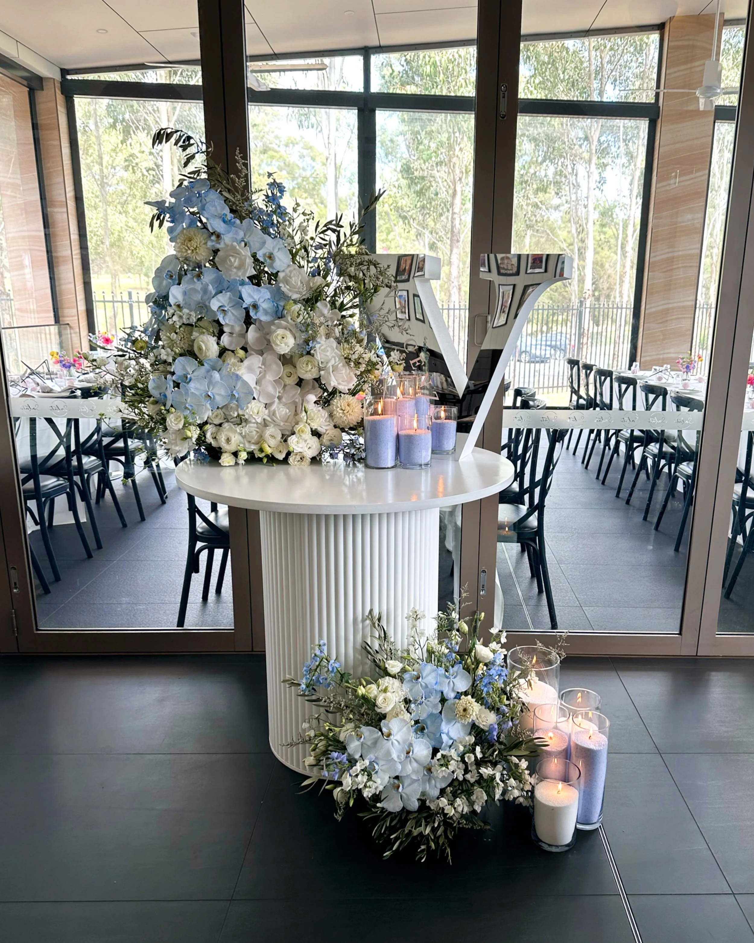 Indoor floral arrangement with white and blue flowers and lit candles on a white table, set against a large window with a view of trees outside.