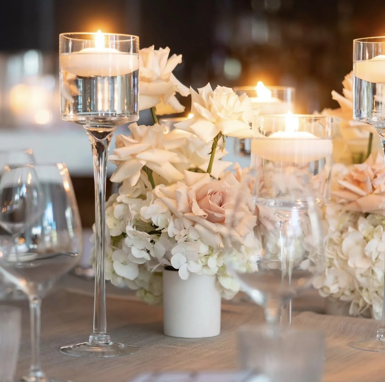 Elegant table centerpiece with white and blush roses, hydrangeas, and floating candles in glass holders.