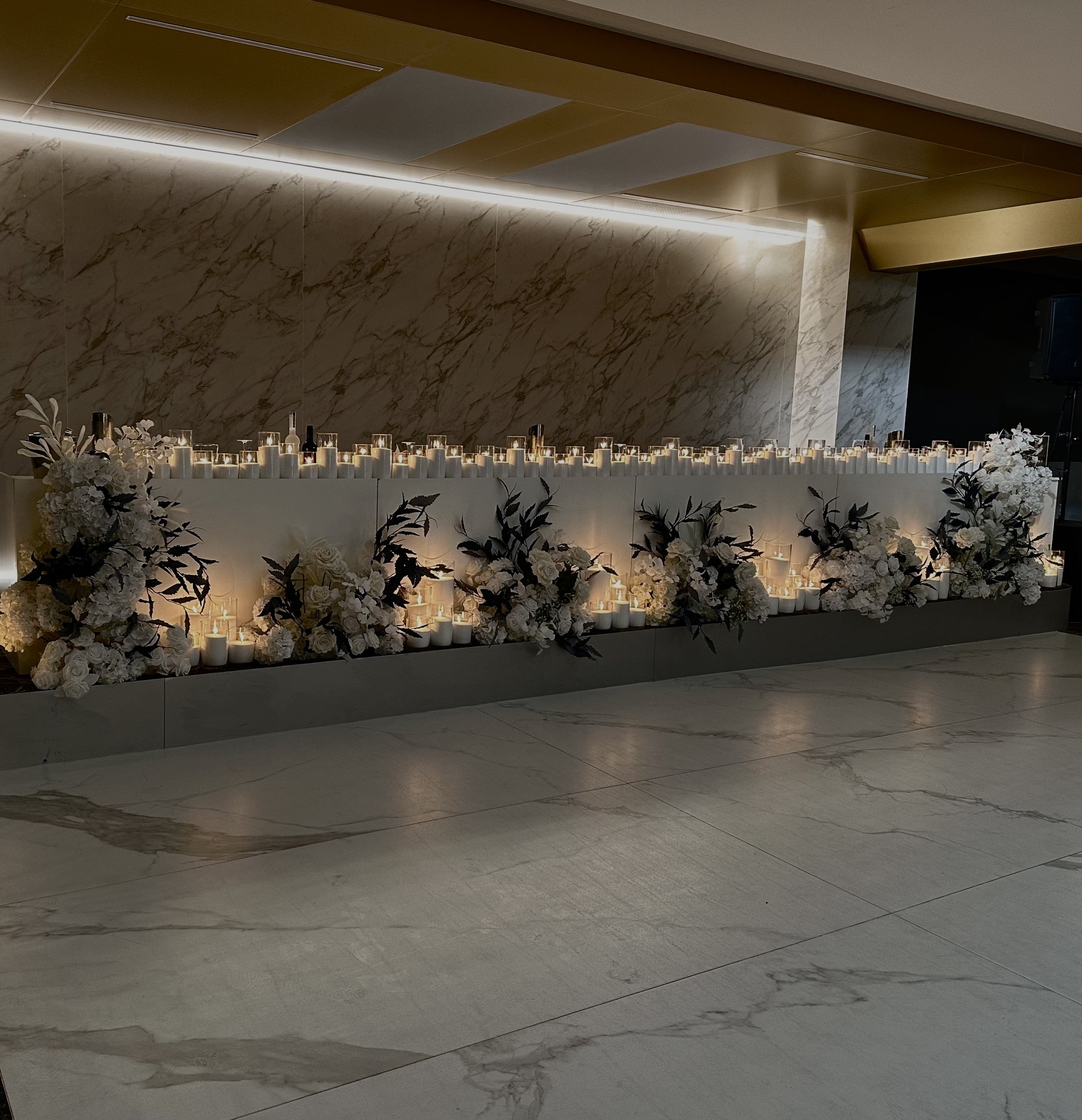A decorative display of numerous white candles with some lit, surrounded by white flowers and greenery, set against a polished marble wall with warm lighting.