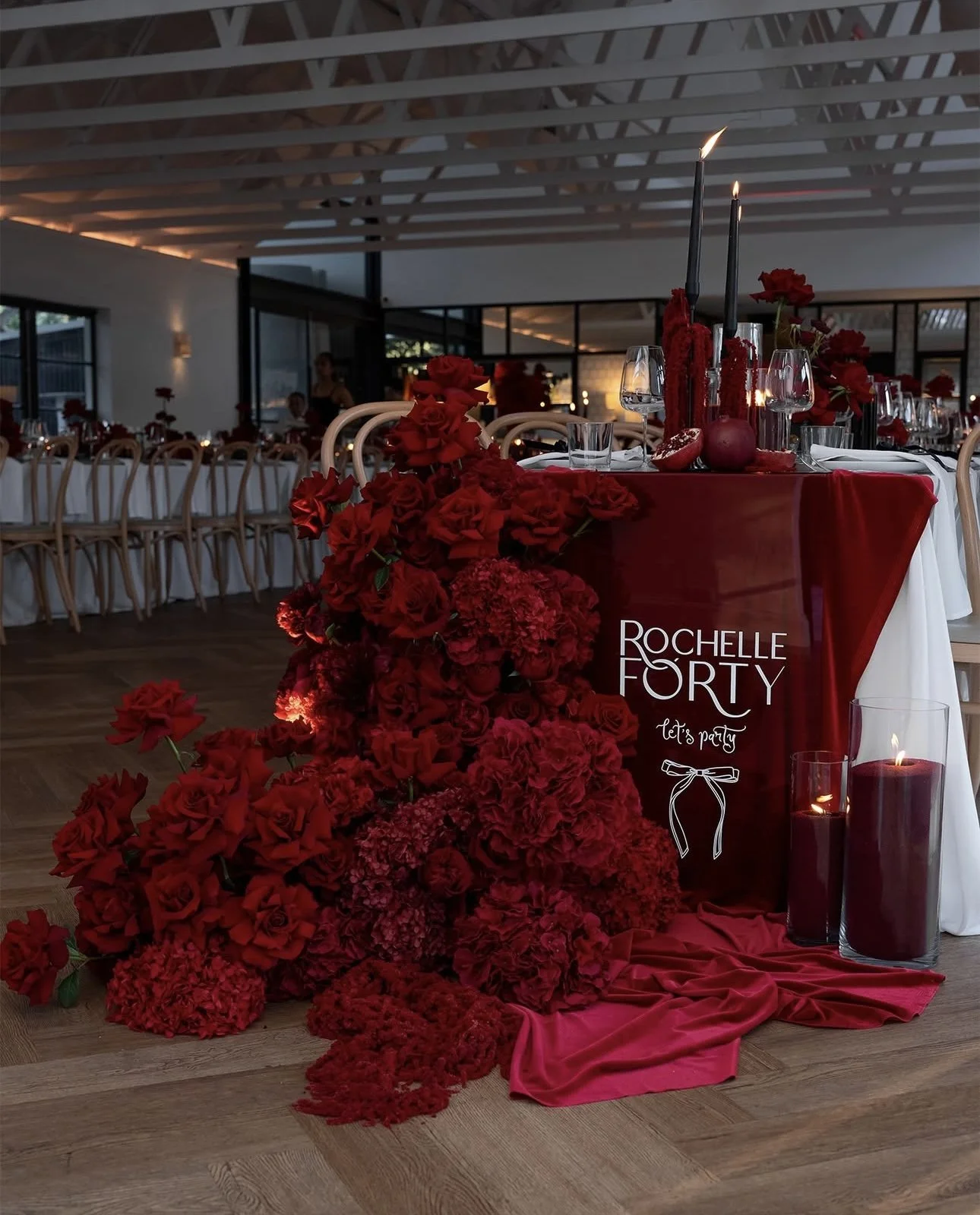 Elegant event table center with red flowers, candles, wine glasses, and a sign reading 'Rochelle Forty let's party' in a modern decorated venue.