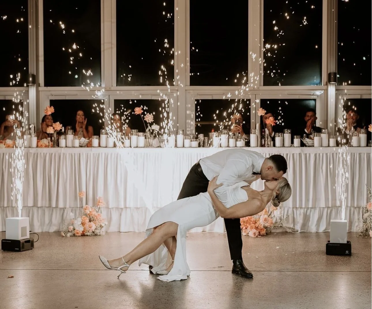 A bride and groom share their first dance at their wedding reception, with guests and sparklers in the background.