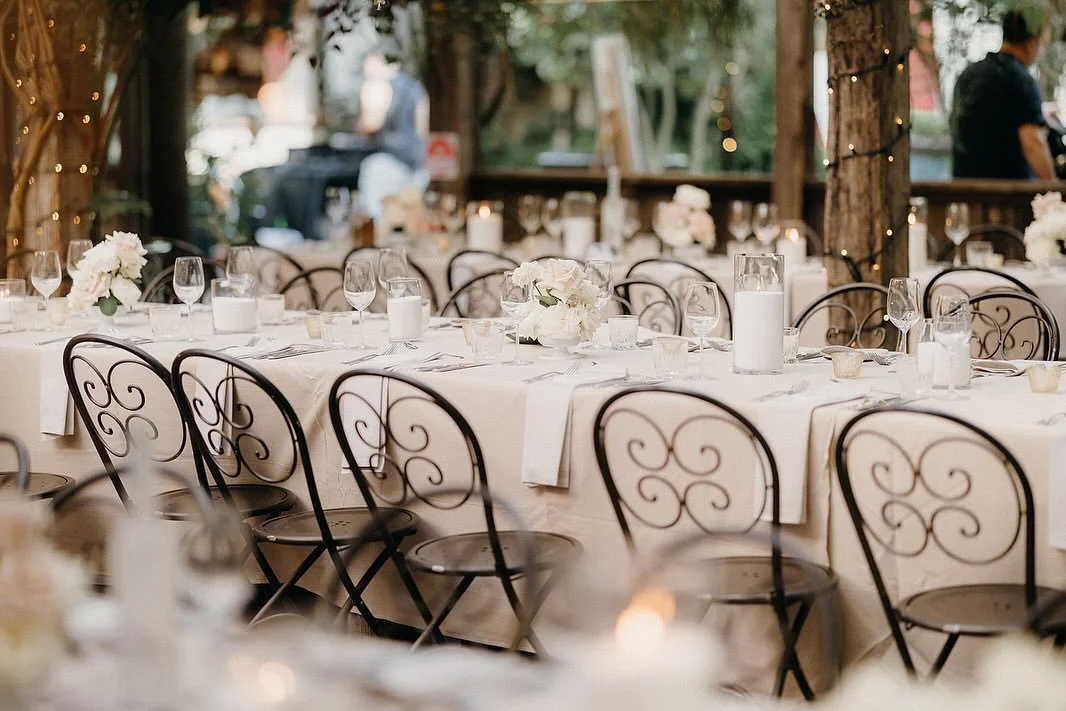 Set dining table with white tablecloth, floral centerpieces, wine glasses, and candles, outdoor setting with string lights and people in the background.