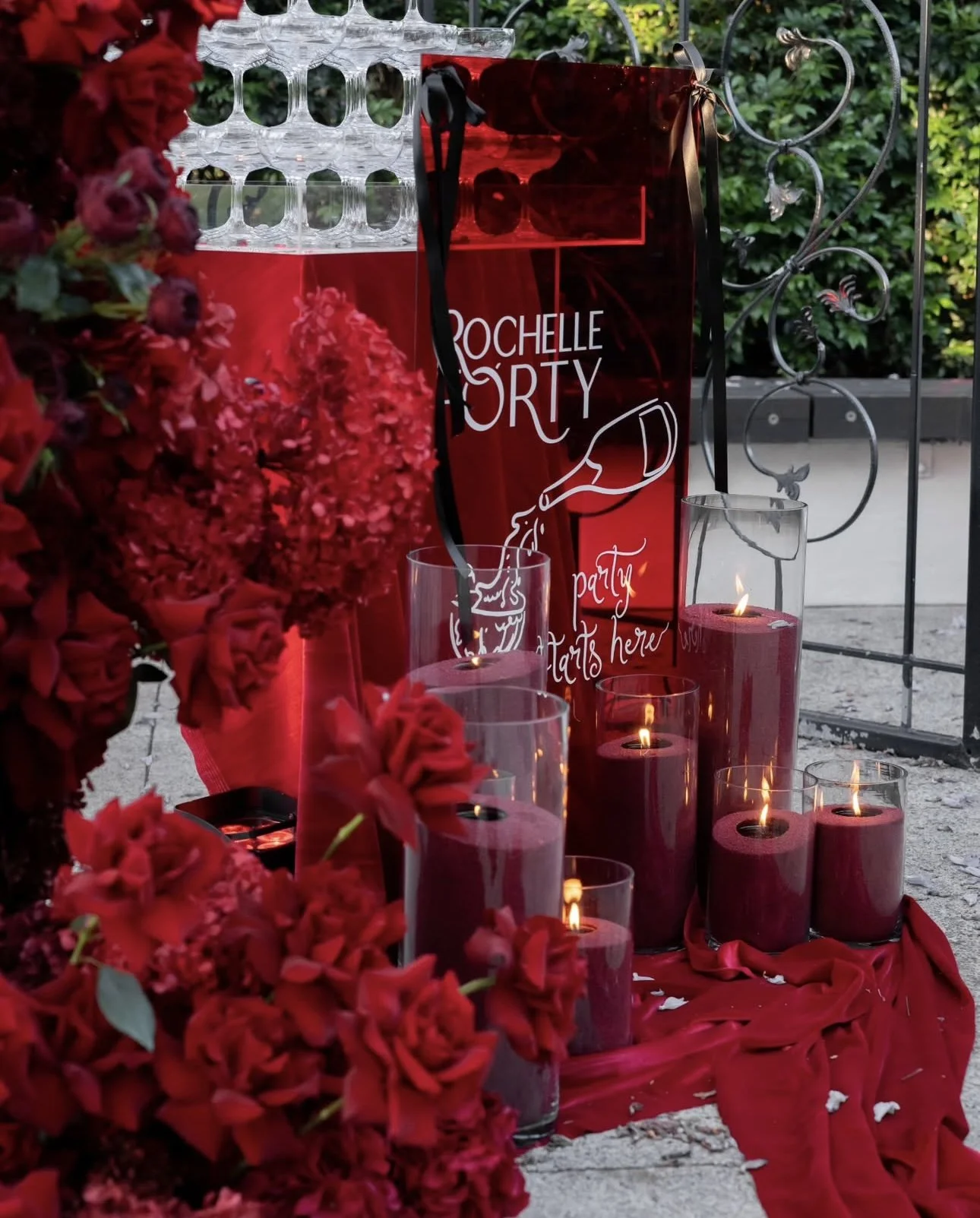 Red floral arrangement and red sand candles in front of a party sign, all set up in front of a champagne tower.