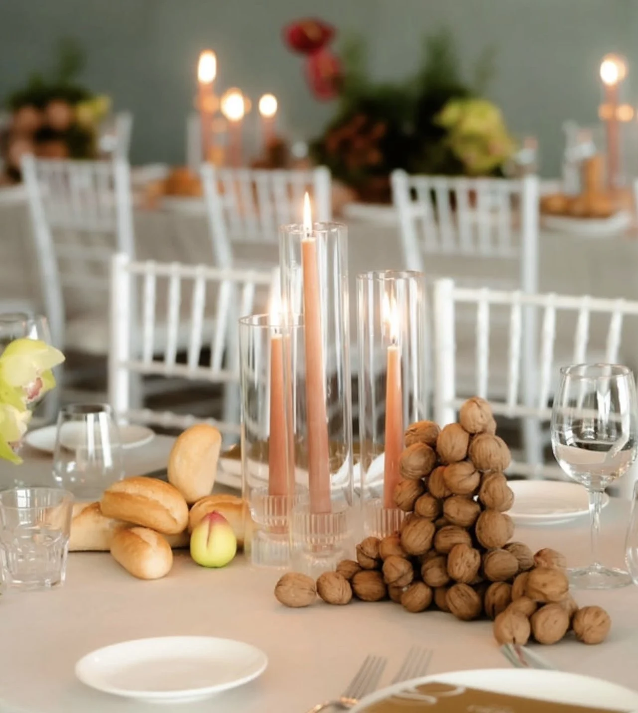 A decorated dining table with candles in glass holders, a pile of walnuts, bread rolls, apples, wine glasses, and tableware, with white chairs and a blurred background with candles and greenery.