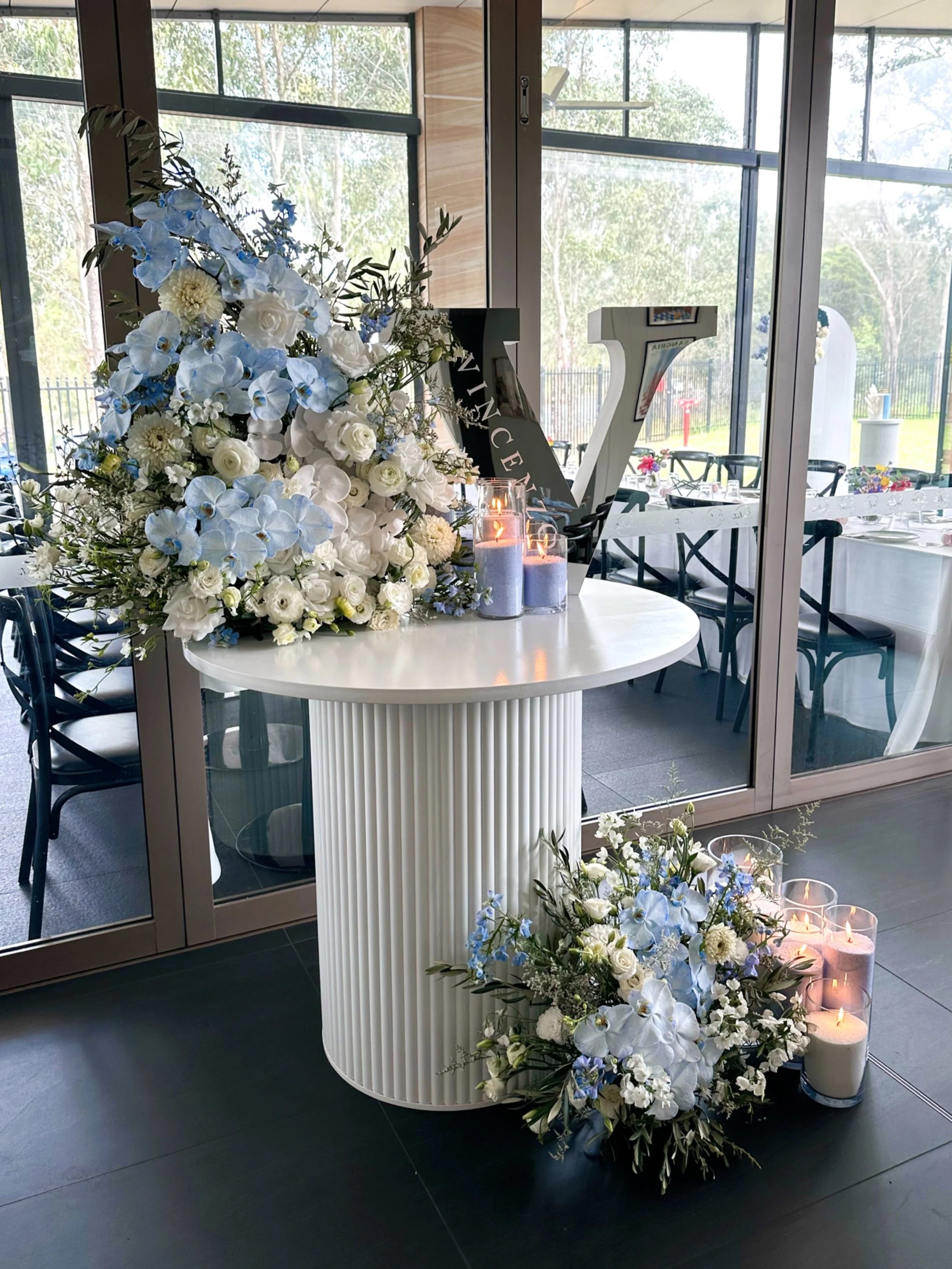 Elegant floral display with white roses, blue orchids, candles, and greenery set on a white pedestal in front of large glass windows at a celebration or wedding venue.