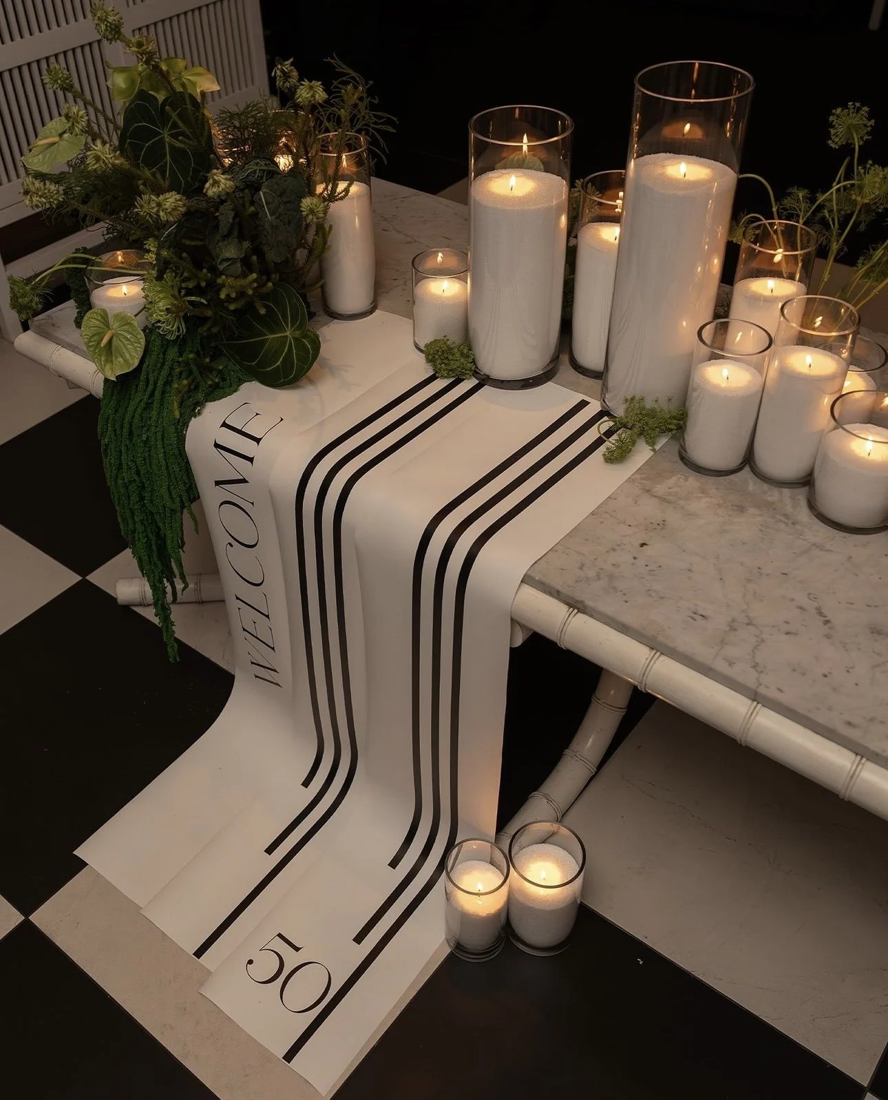 Decorative table display with white candles in glass containers, greenery, and a designer fabric runner with black lines and 'NEVE COME 50' printed on it, set on a marble top table.