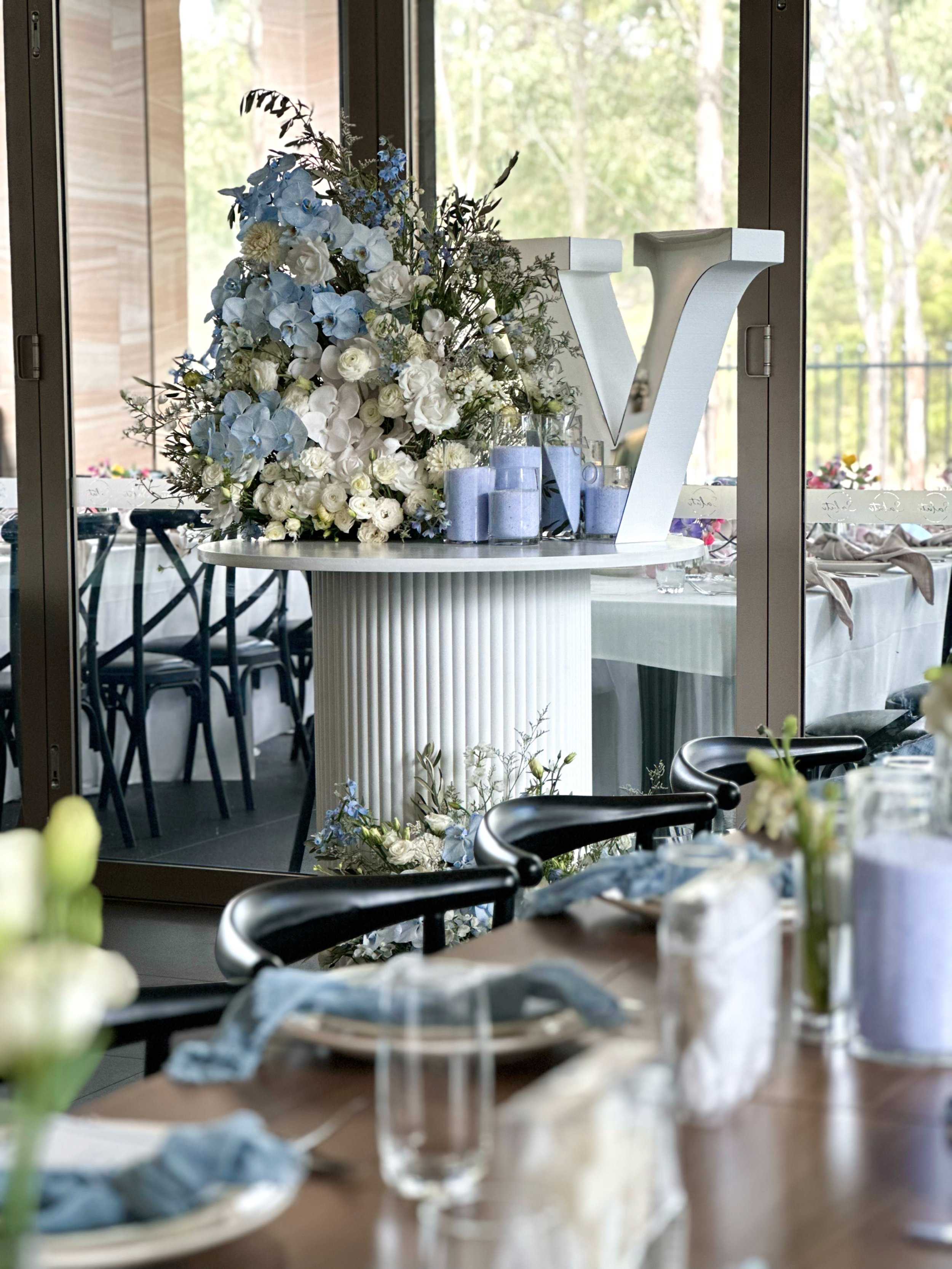 An indoor event space decorated with a large floral arrangement of white and blue flowers and a white decorative letter V on a round table. The space has large glass doors with a view of trees outside.