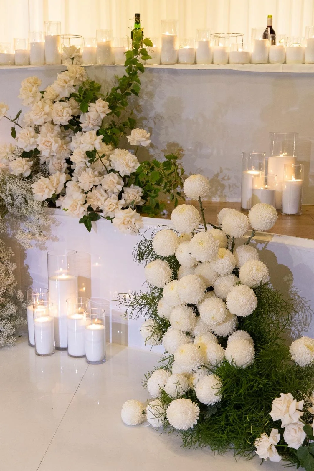 White floral arrangement with candles and glass containers on a white surface, likely part of a wedding or event decoration.