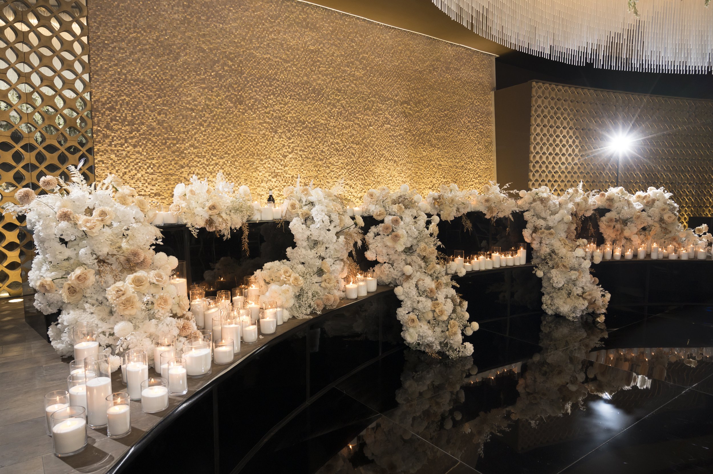 Elegant event setup featuring a large arrangement of white and beige flowers, surrounded by lit candles on the floor and tables, against a textured gold backdrop and geometric wall designs.