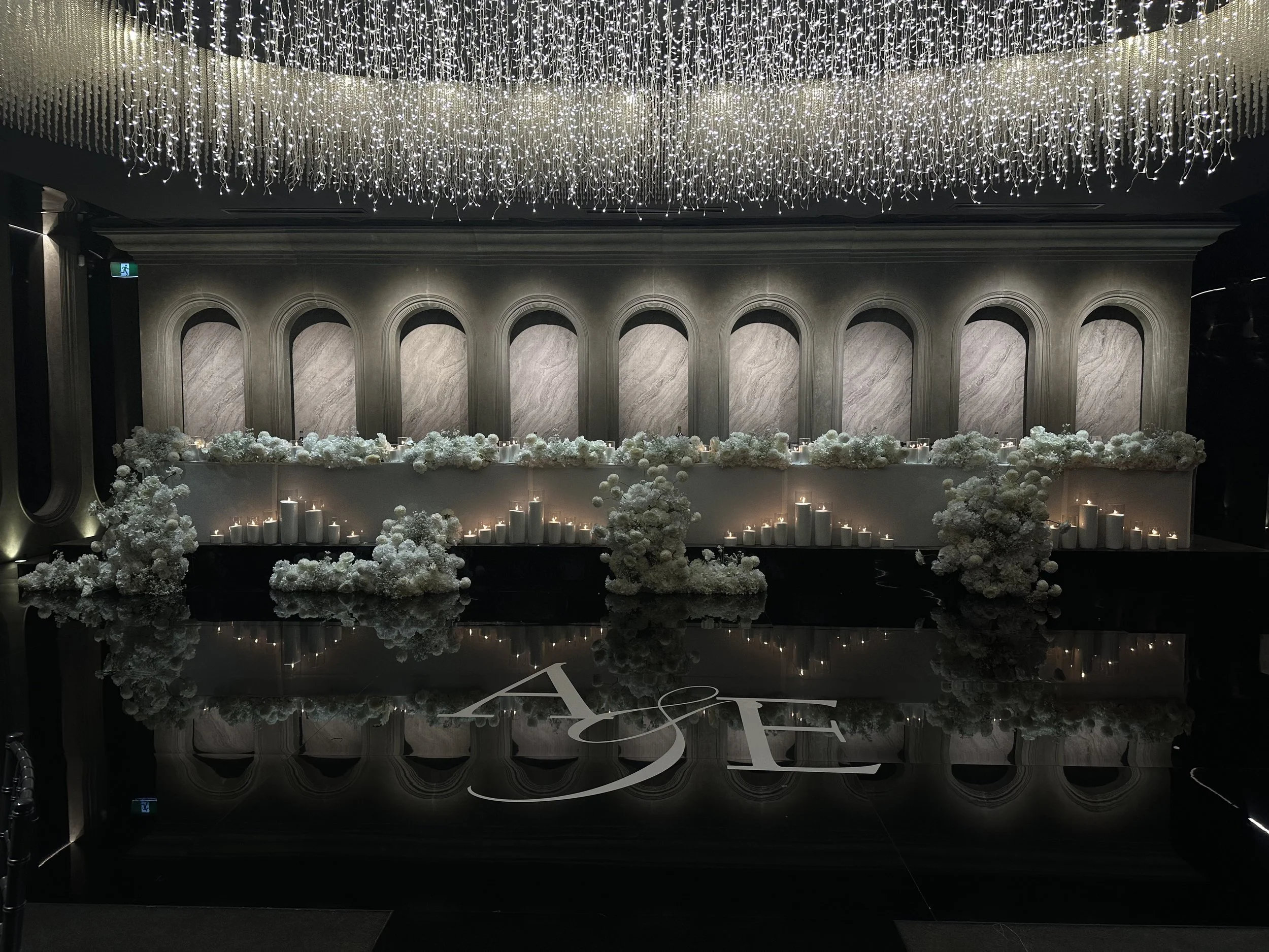 Elegant indoor event space decorated with white flowers, candles, and hanging string lights on the ceiling.