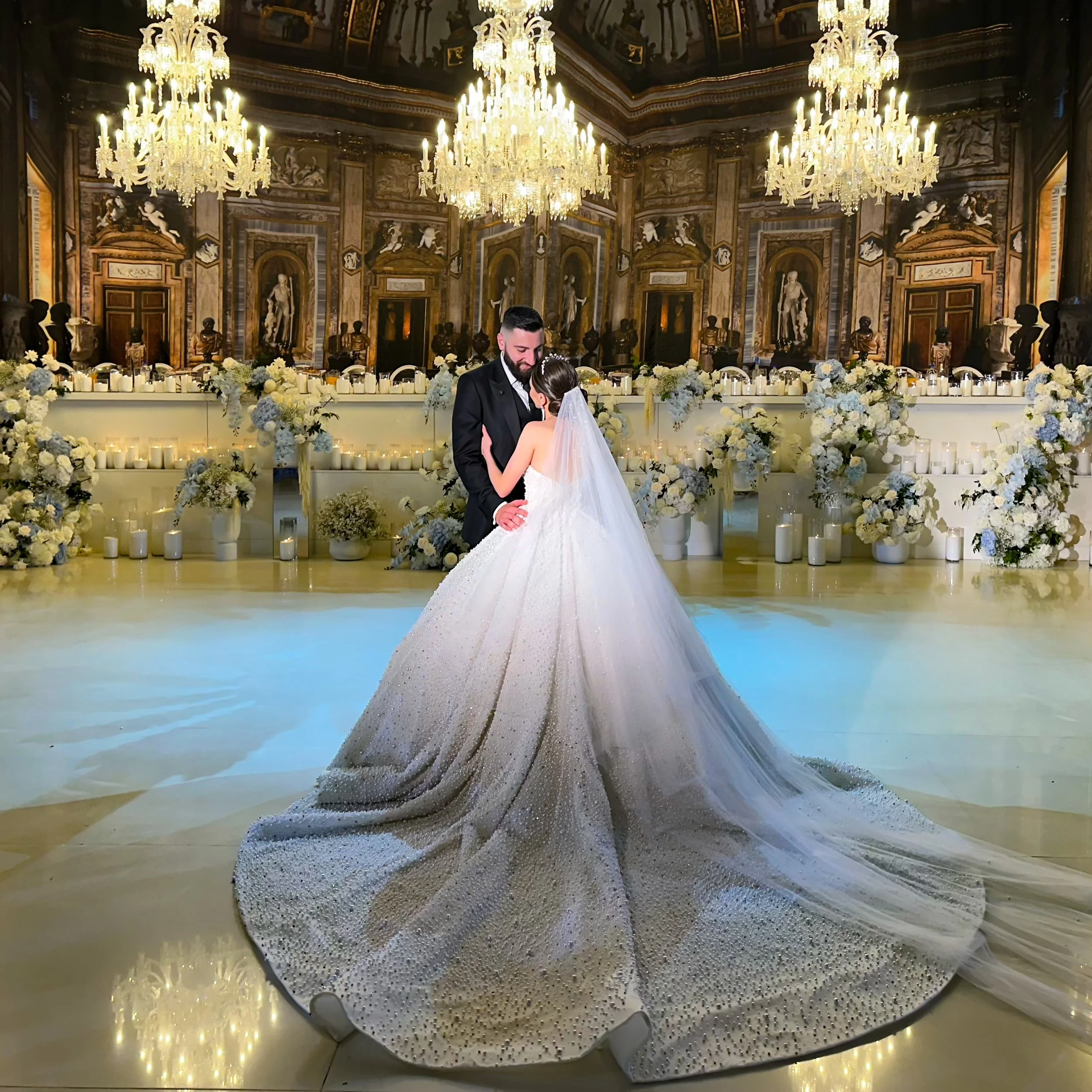 A couple dancing at a wedding reception in an opulent hall with large chandeliers, classical statues, and floral decorations.